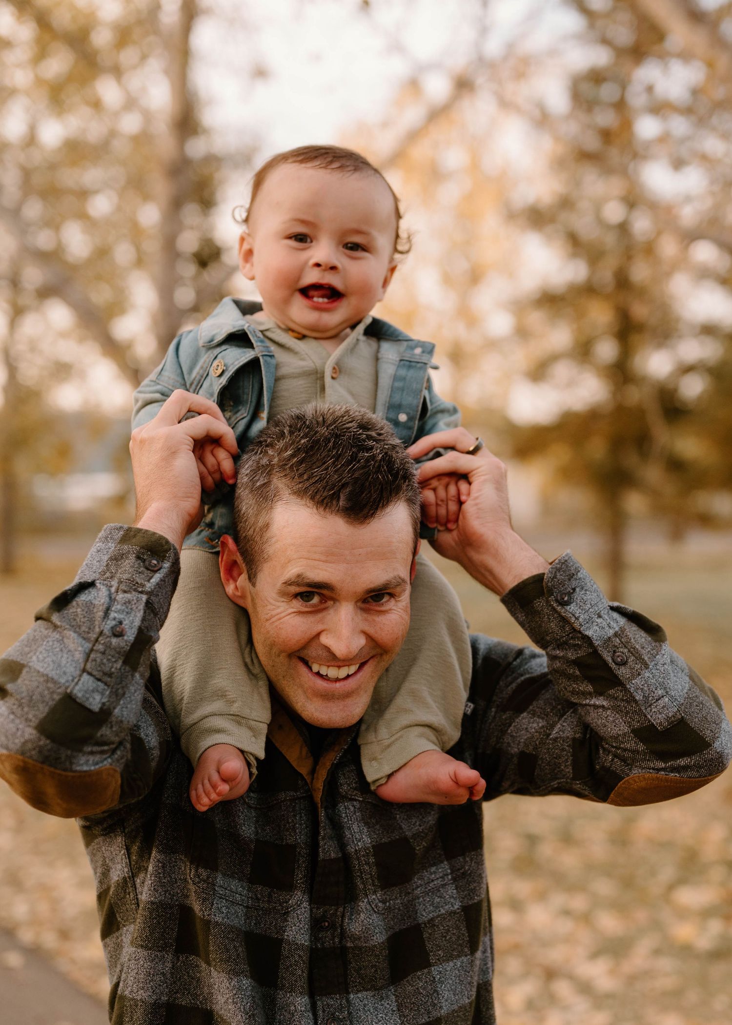 Cheerful father smiling with happy baby sitting on his shoulders in golden autumn park setting.
