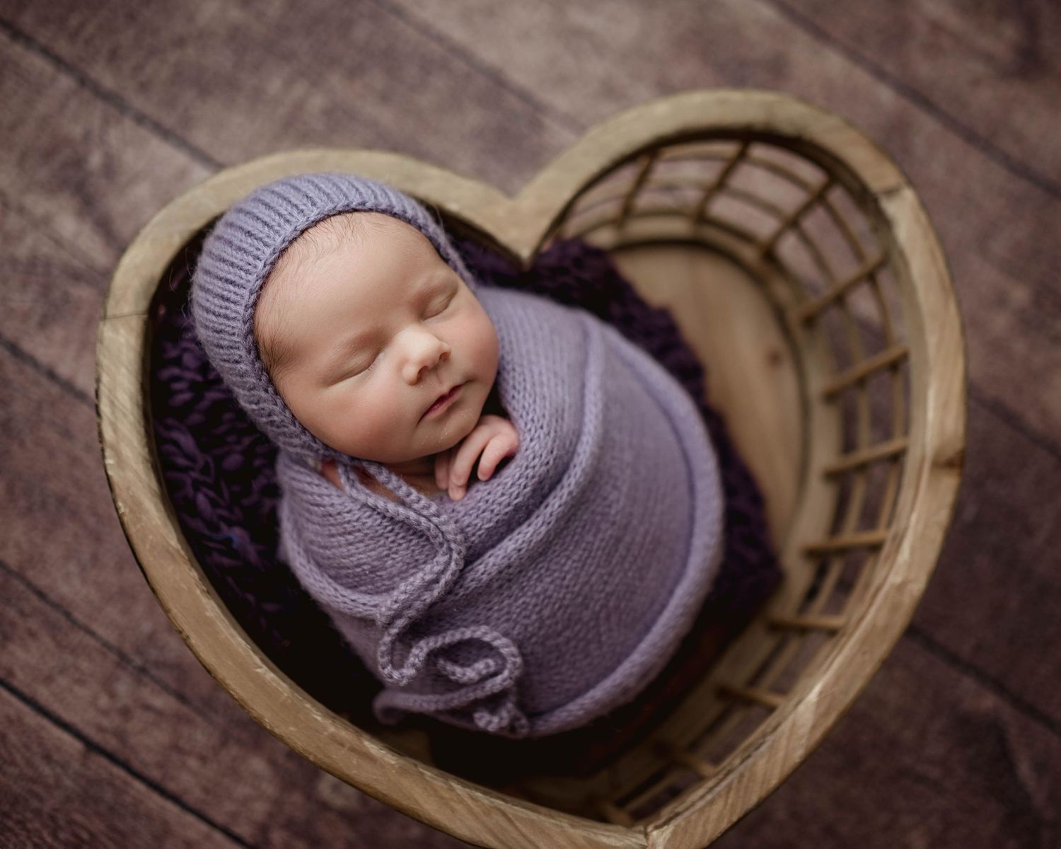 Newborn baby wrapped in purple knit swaddle sleeping in heart-shaped wooden basket with matching bonnet.