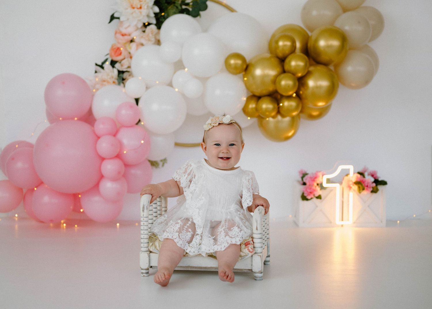 Baby girl in white lace dress sits on bench at first birthday with pink, white, and gold balloon garland.