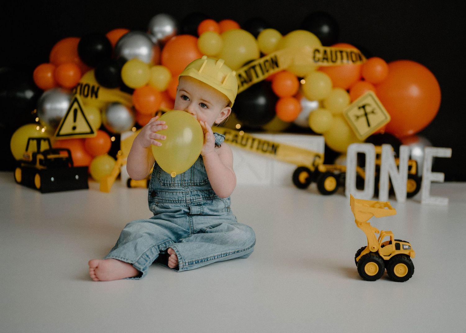 Baby boy in denim overalls and hard hat plays with yellow balloon at construction-themed first birthday.