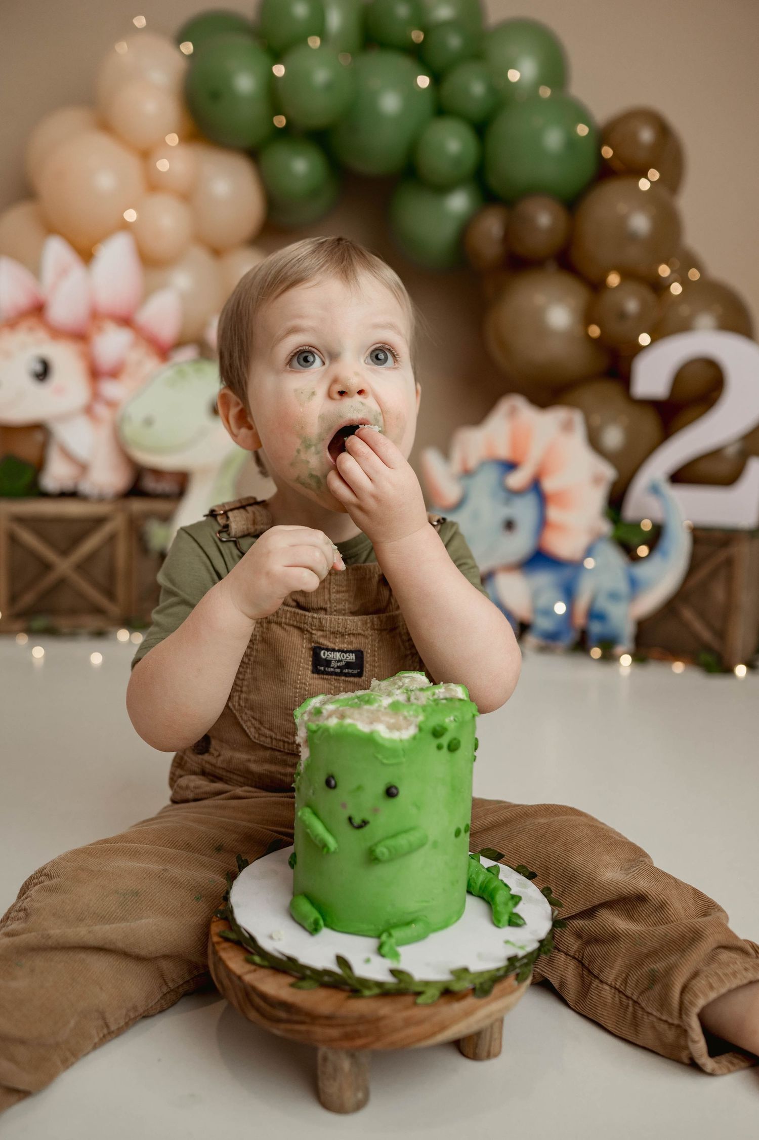 Toddler boy in overalls eats green dinosaur birthday cake with hands at a dino-themed cake smash.