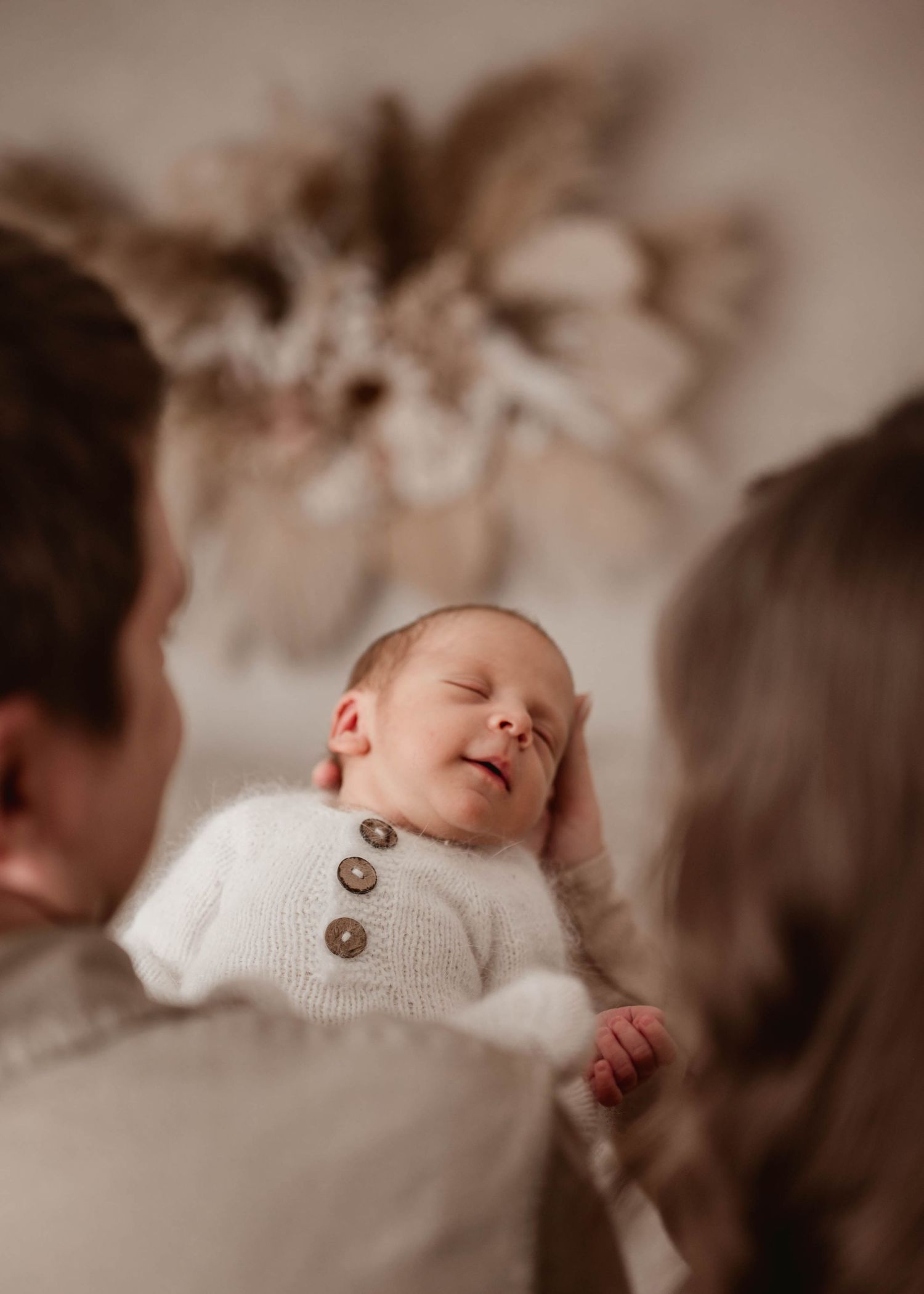 Smiling newborn in white knit outfit held by parents with boho floral decor in background.