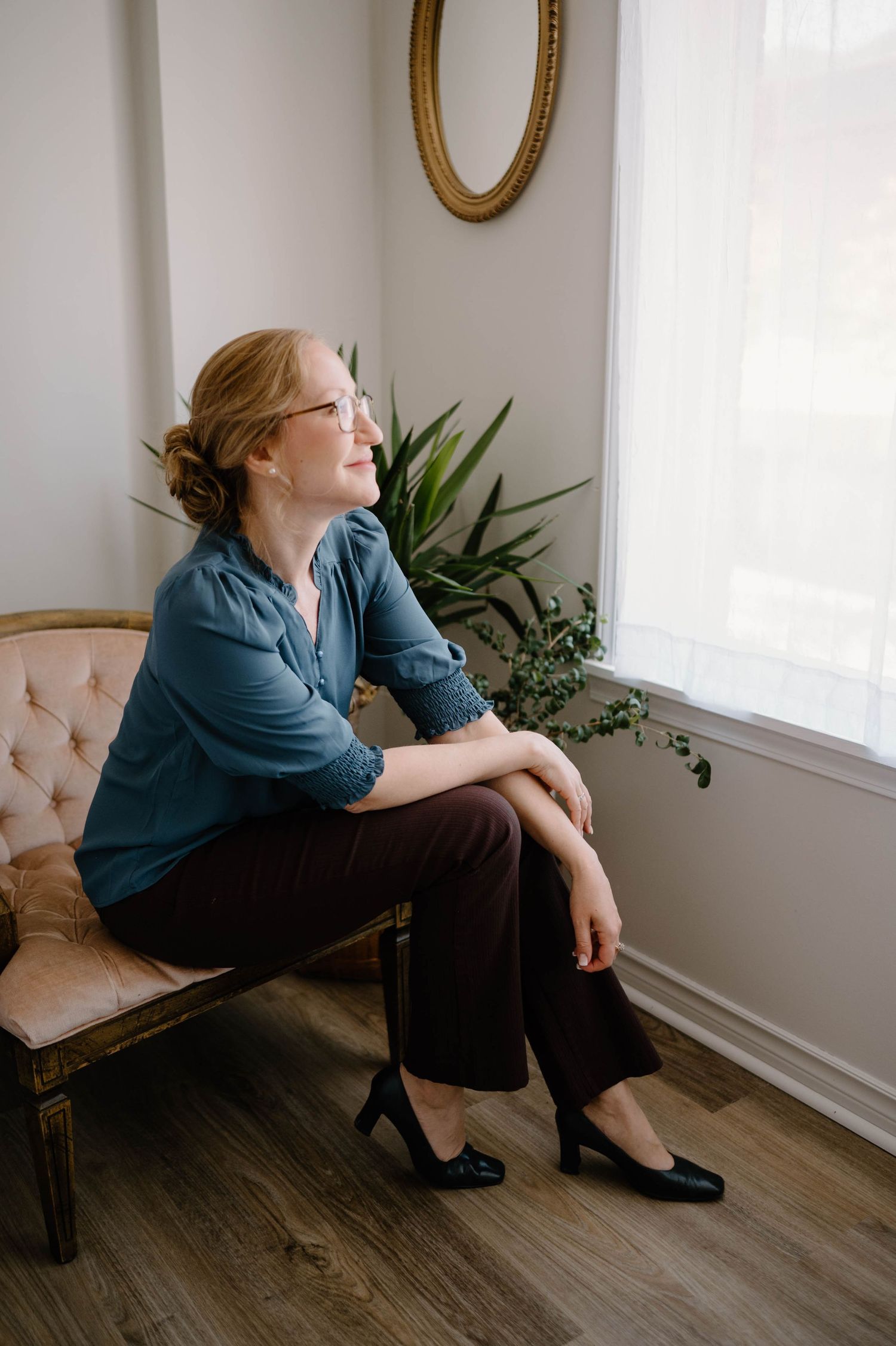 Woman in teal blouse and brown pants sitting on vintage bench, gazing out bright window.