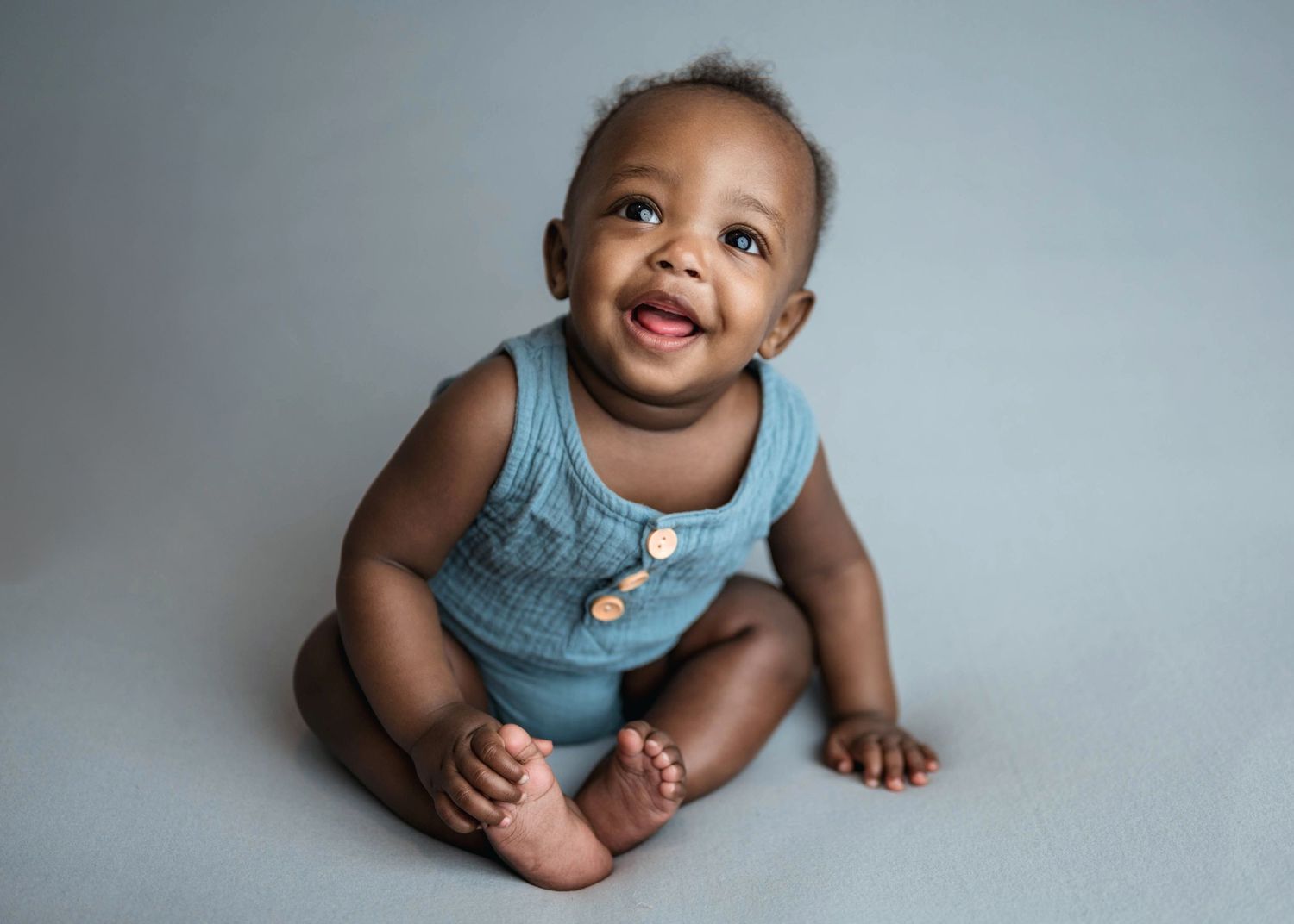 Happy smiling baby boy sitting upright wearing blue romper against light gray studio background.