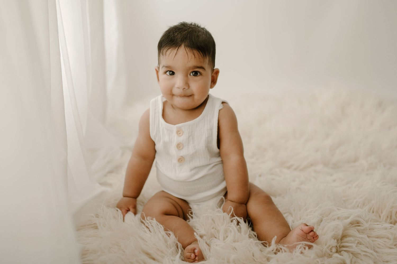 Adorable baby in a white button romper sitting on a fluffy cream rug in a softly lit white studio.