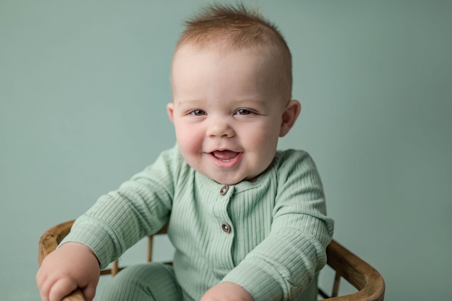 Smiling baby boy wearing sage green ribbed romper seated in a wicker basket on teal background.
