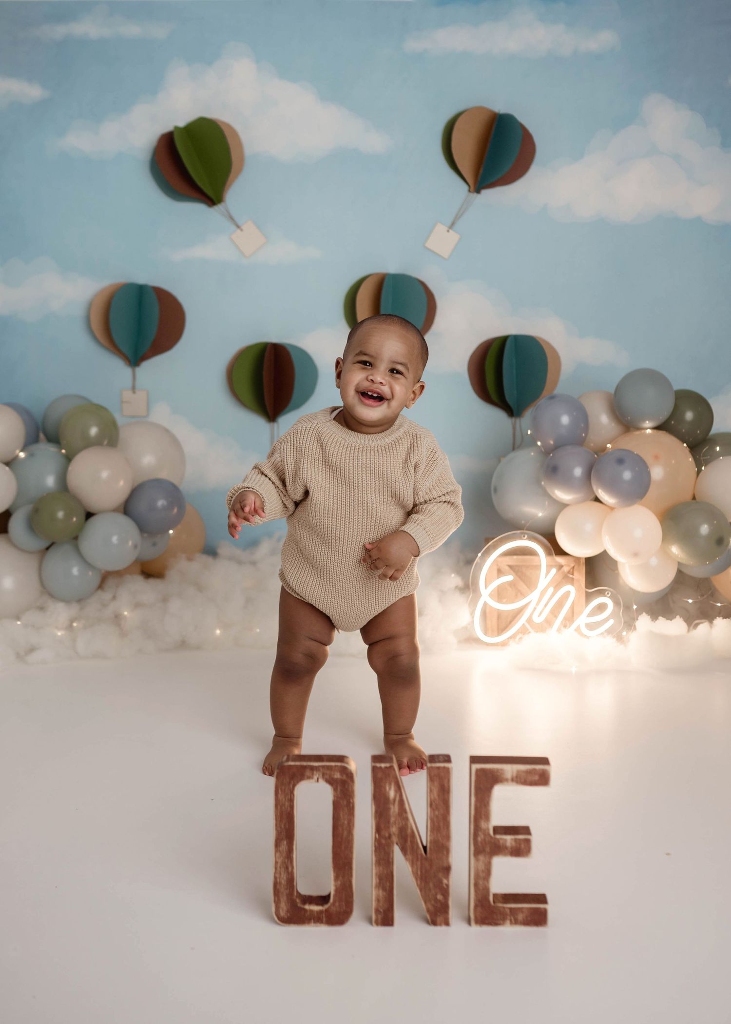 Joyful baby standing over wooden ONE letters at a hot air balloon themed first birthday cake smash photo session.