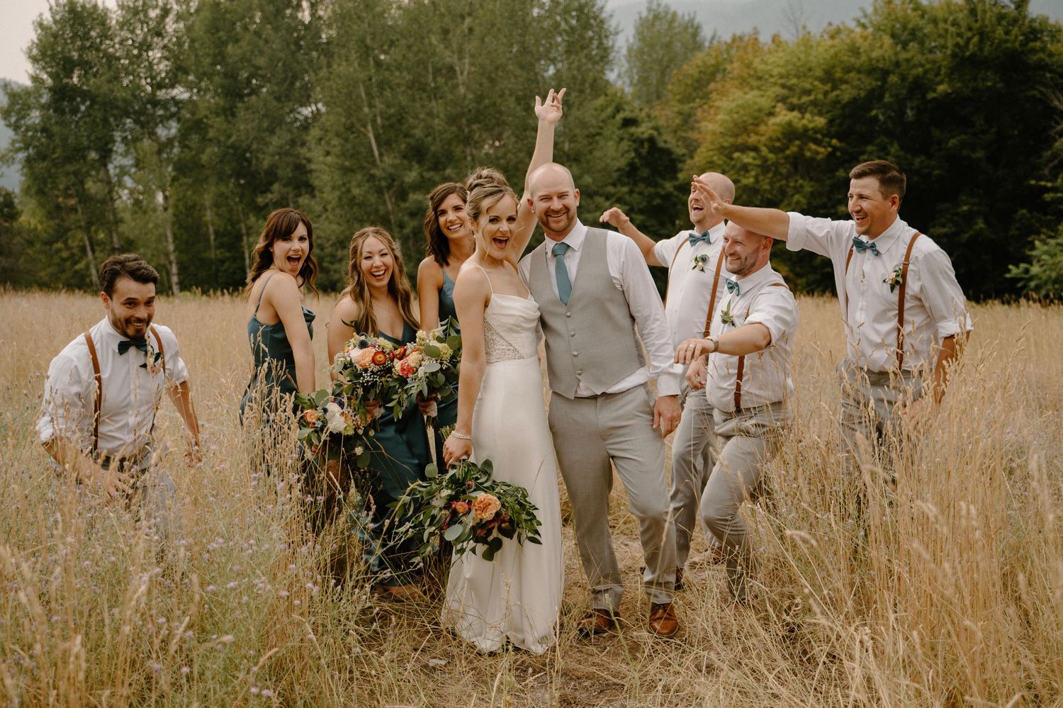 Joyful wedding party celebrating in a golden field with the bride holding a colorful floral bouquet.