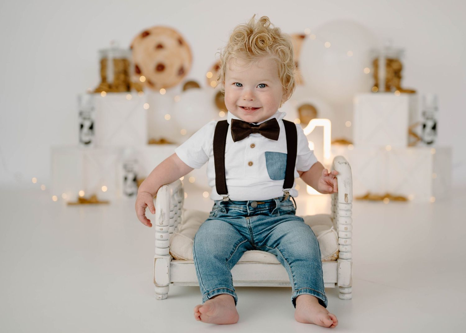 Smiling baby boy in suspenders and jeans sits on white chair at cookies and milk first birthday session.