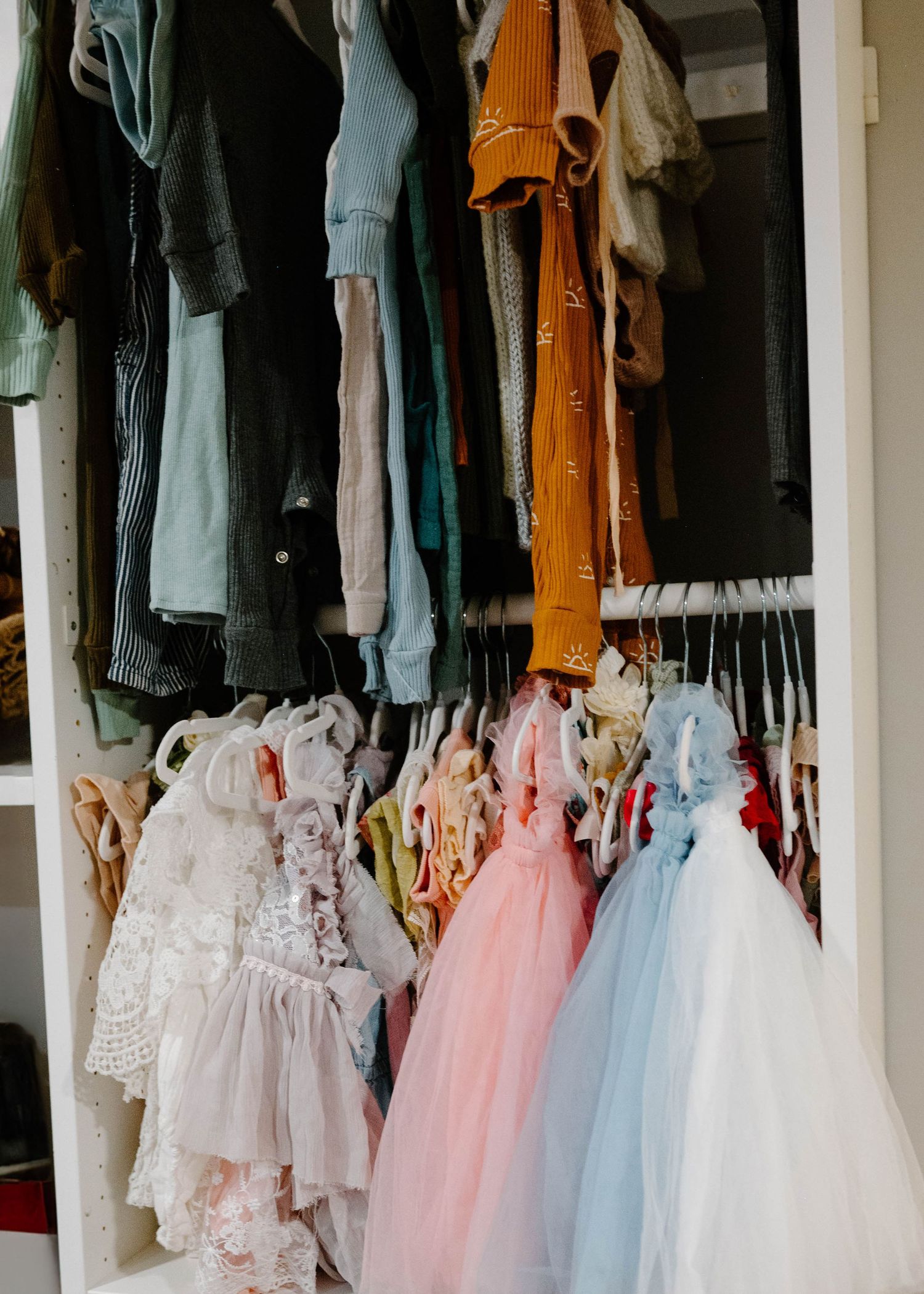 Colorful children's dresses and clothing hanging on racks inside a white wardrobe storage unit.