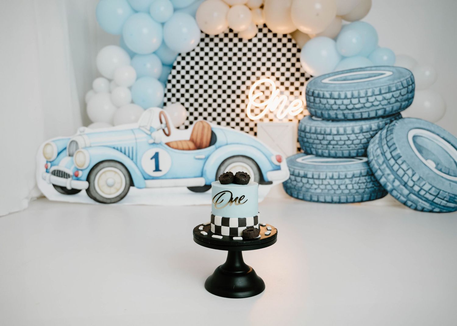 A vintage blue race car prop and stacked tire decorations with a checkered cake displayed at a first birthday photo session.
