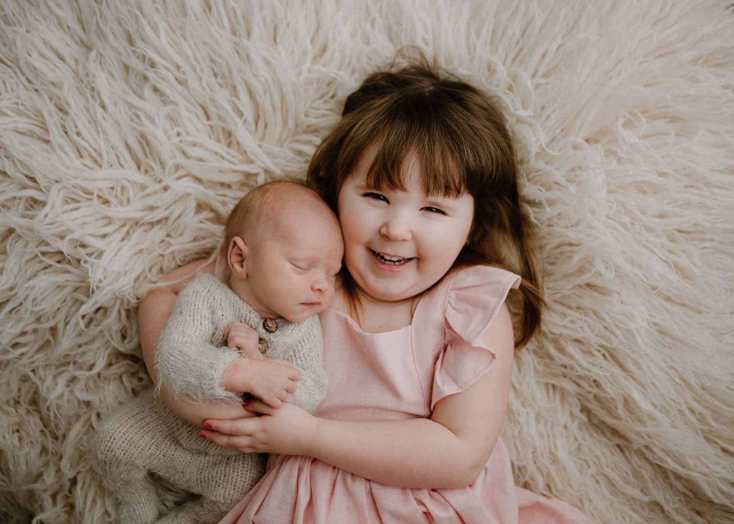 Smiling toddler girl in pink dress cuddles sleeping newborn sibling on a fluffy cream fur rug.