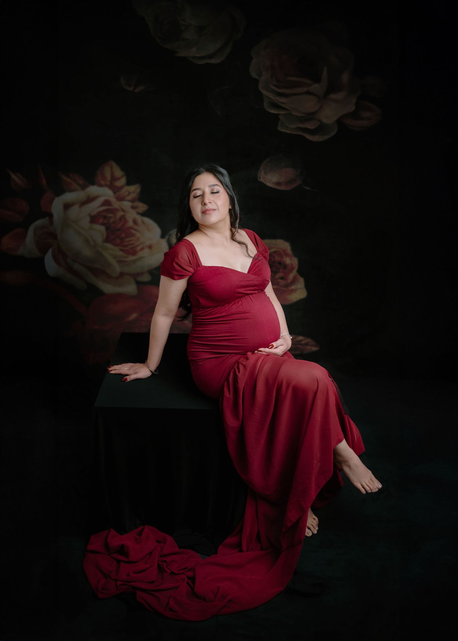 Pregnant woman in elegant red gown seated on black cube against dark floral backdrop.