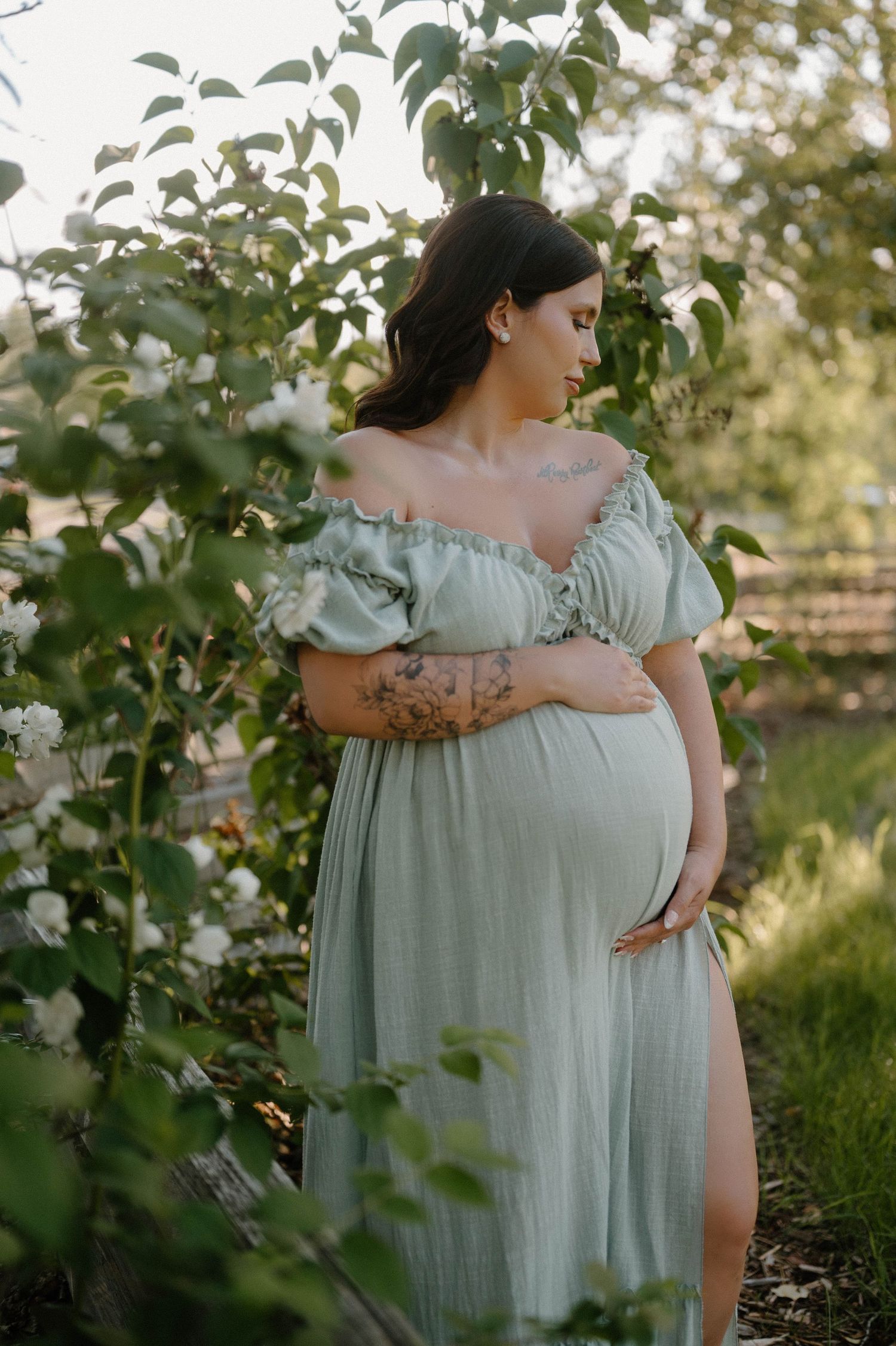 Pregnant woman in sage green off-shoulder dress cradling baby bump among blooming white flowers outdoors.