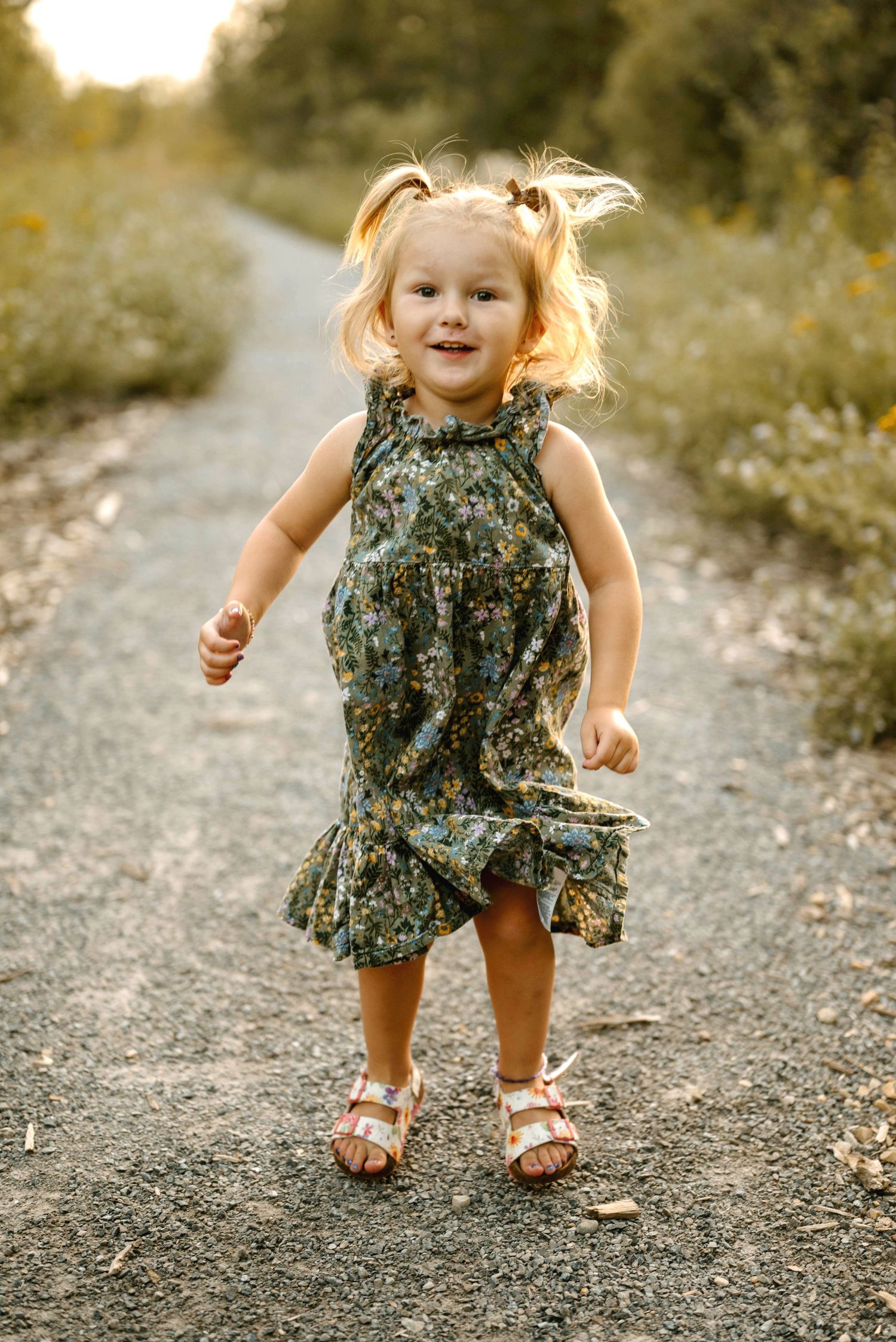 Toddler girl in floral dress running on a sunlit dirt path surrounded by greenery.