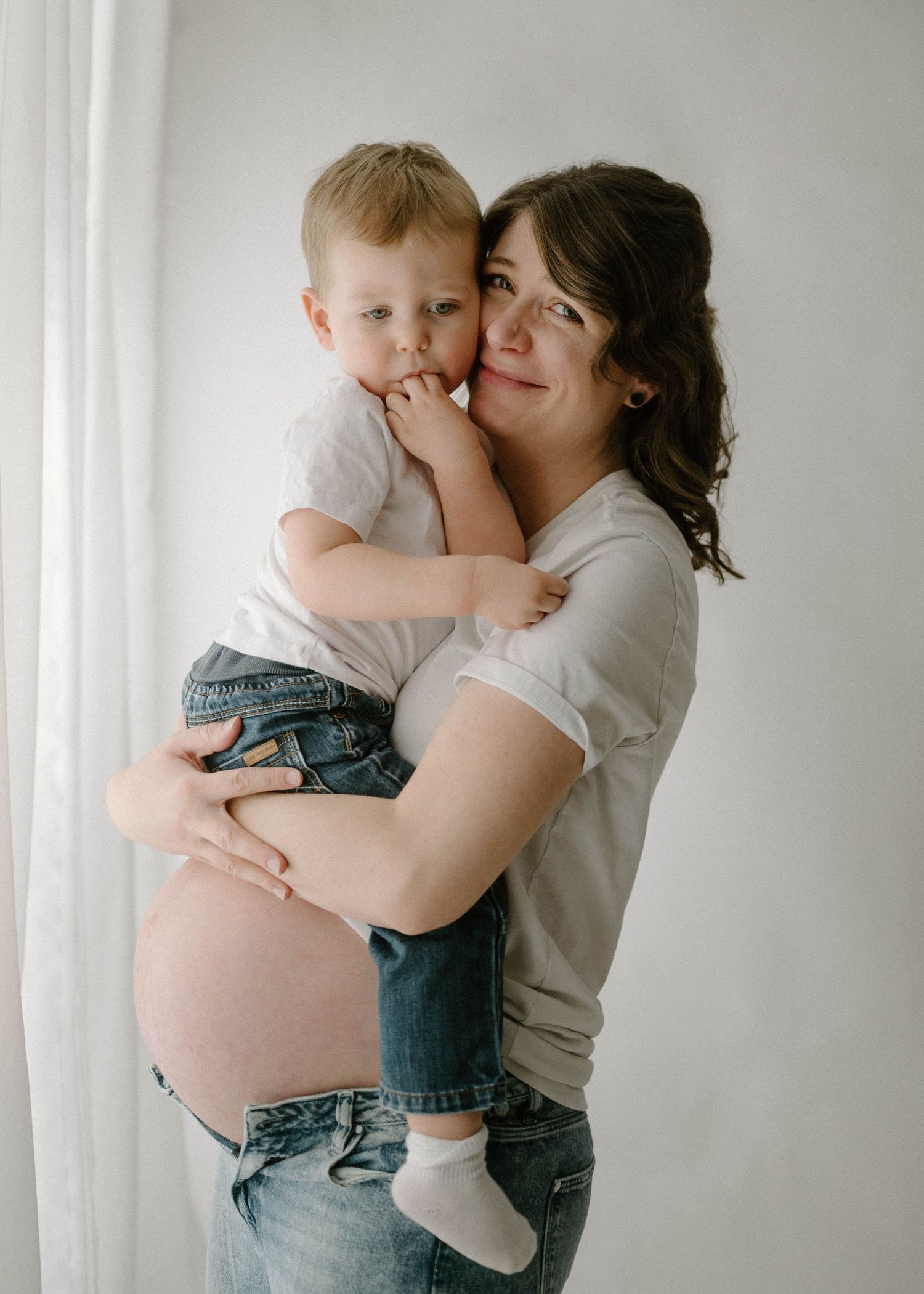 Pregnant woman in white tee holds smiling toddler son close near a bright window curtain.