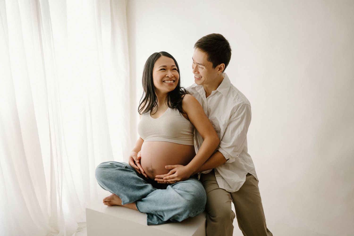 Happy expectant couple sitting together on white cube, woman cradling baby bump, smiling warmly.