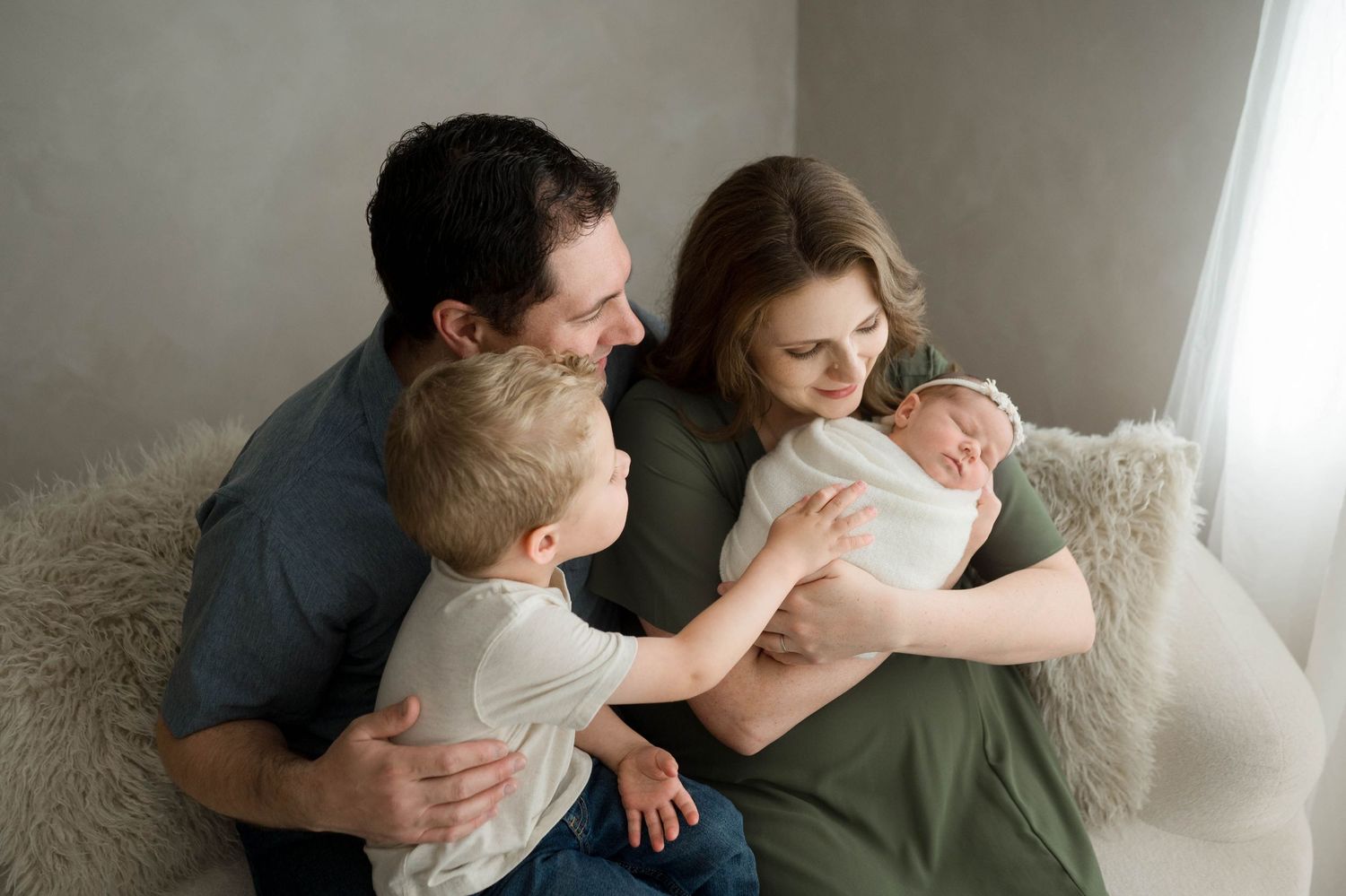Family of four with newborn baby, parents and toddler son sitting together on a couch in a bright studio.