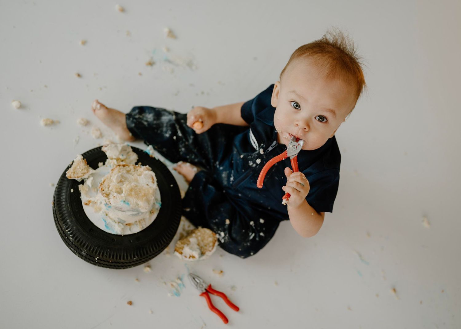Overhead view of baby boy covered in cake eating with spoon at garage-themed first birthday smash session.
