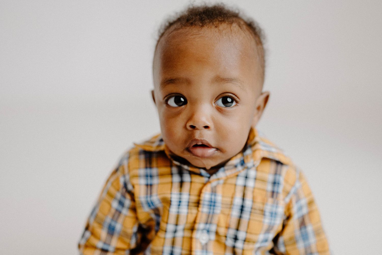 Close-up portrait of a cute Black toddler boy in a yellow plaid shirt looking at the camera.