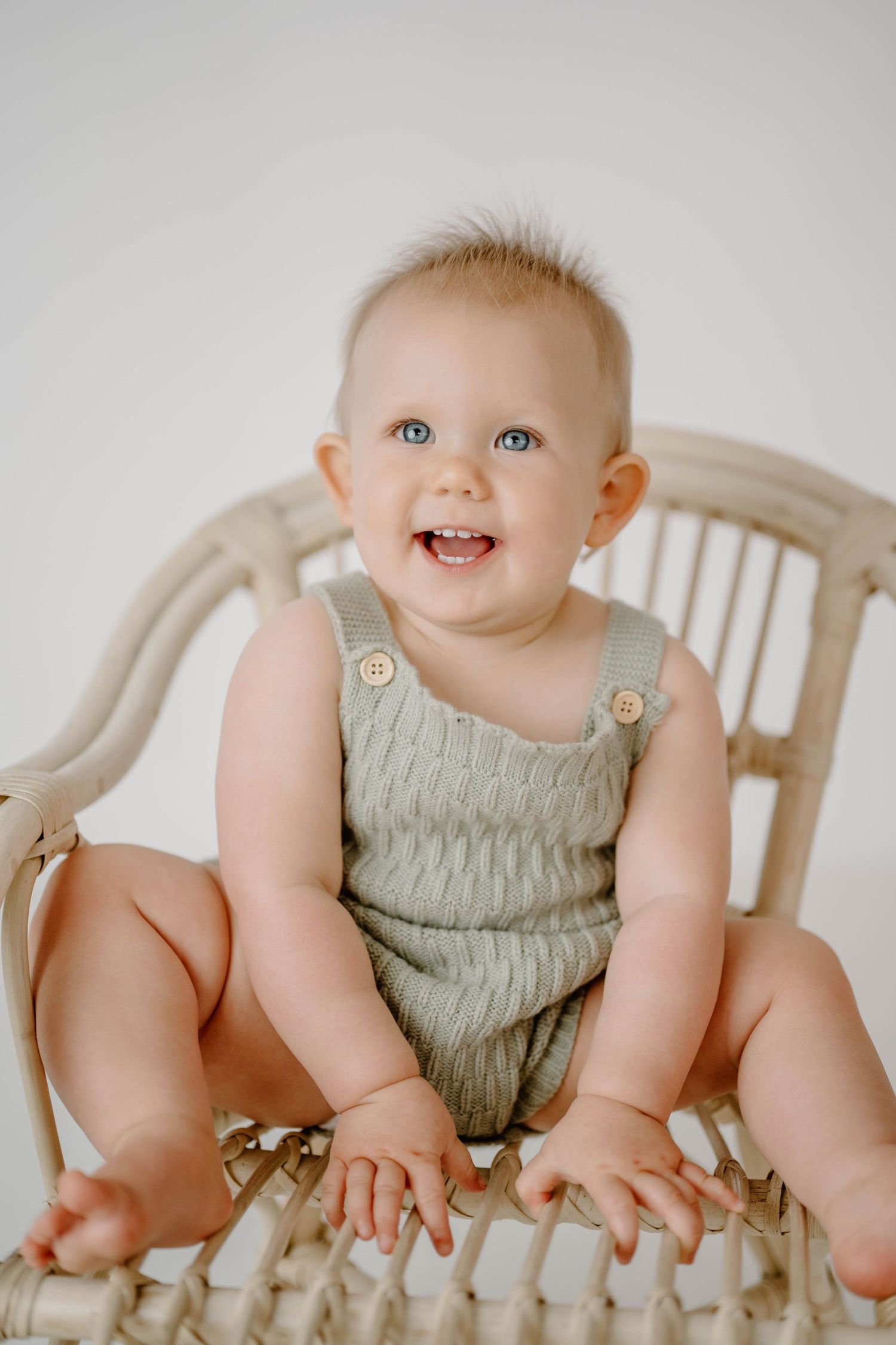Cheerful blue-eyed baby in sage green knit romper sitting in rattan chair smiling.
