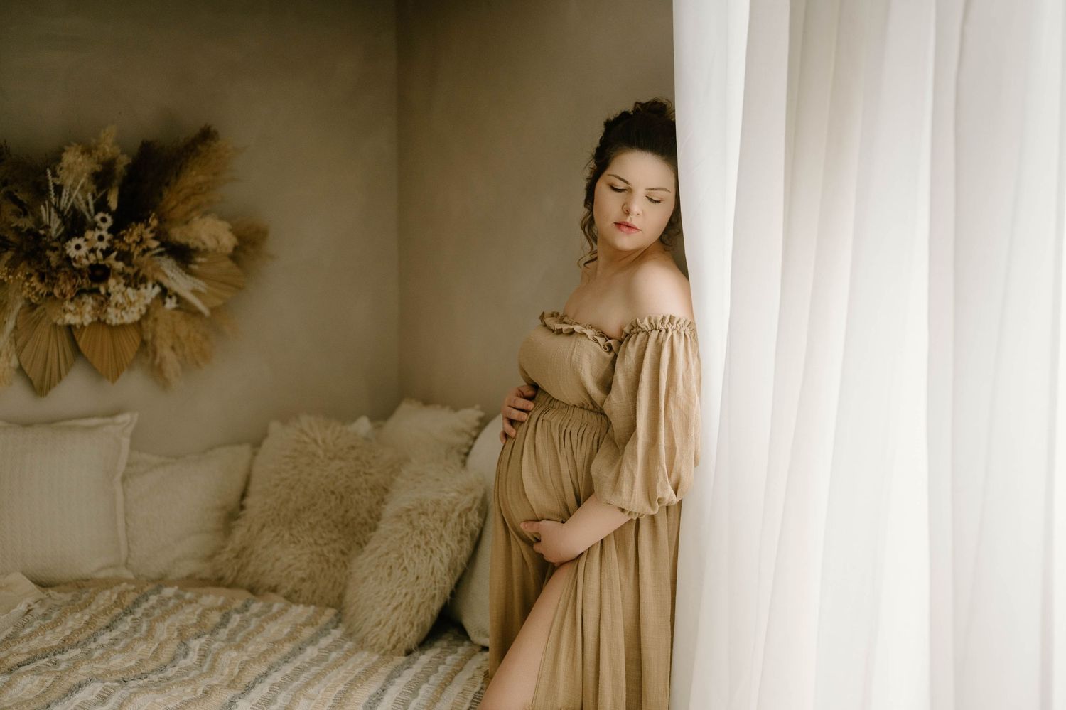 Pregnant woman in golden off-shoulder maternity gown standing by white curtains in boho bedroom.