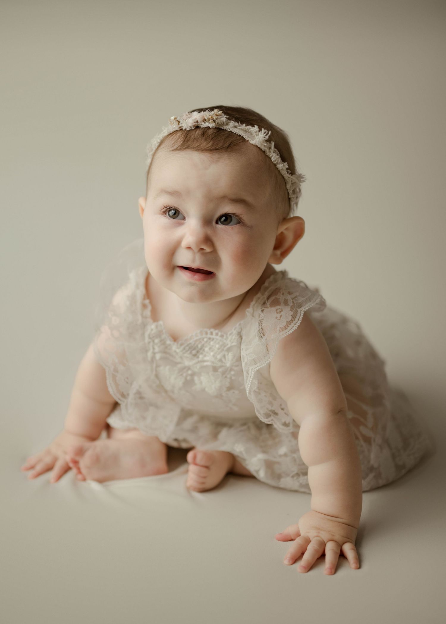 Happy baby girl in ivory lace romper and headband crawling on neutral studio backdrop during milestone photo session.