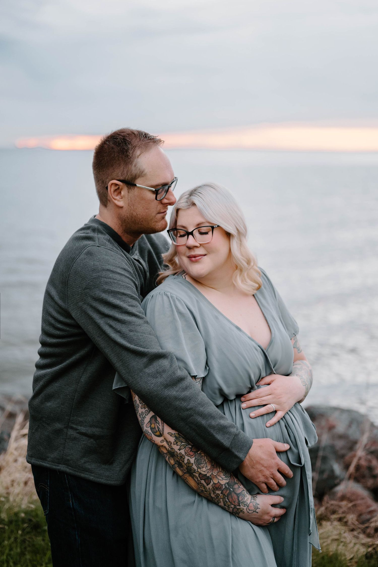 Couple embracing during maternity photoshoot by rocky ocean shore at sunset, man with tattoos hugging pregnant woman in teal dress.
