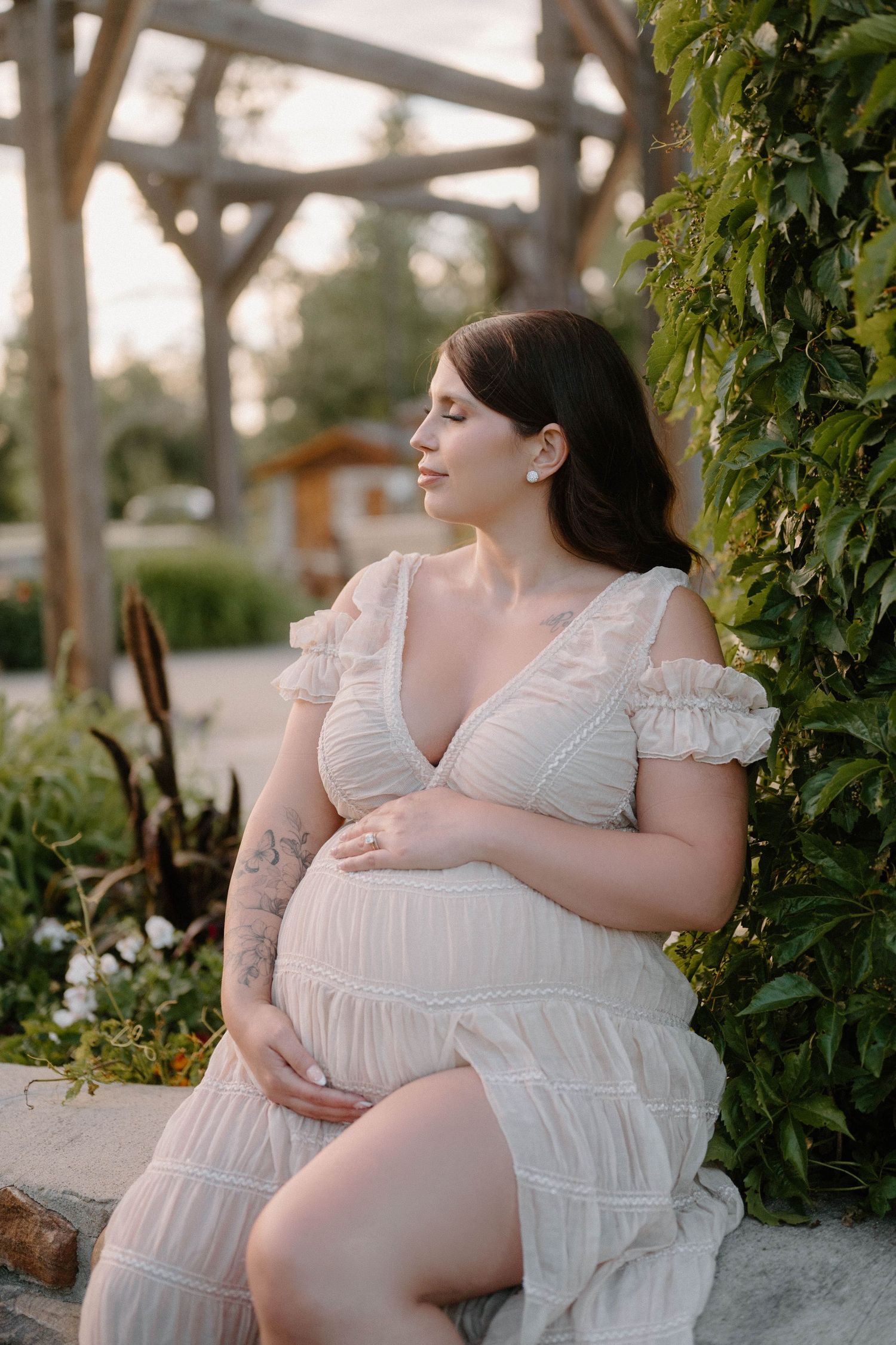 Pregnant woman in white lace dress sitting outdoors among green plants, cradling baby bump with eyes closed.