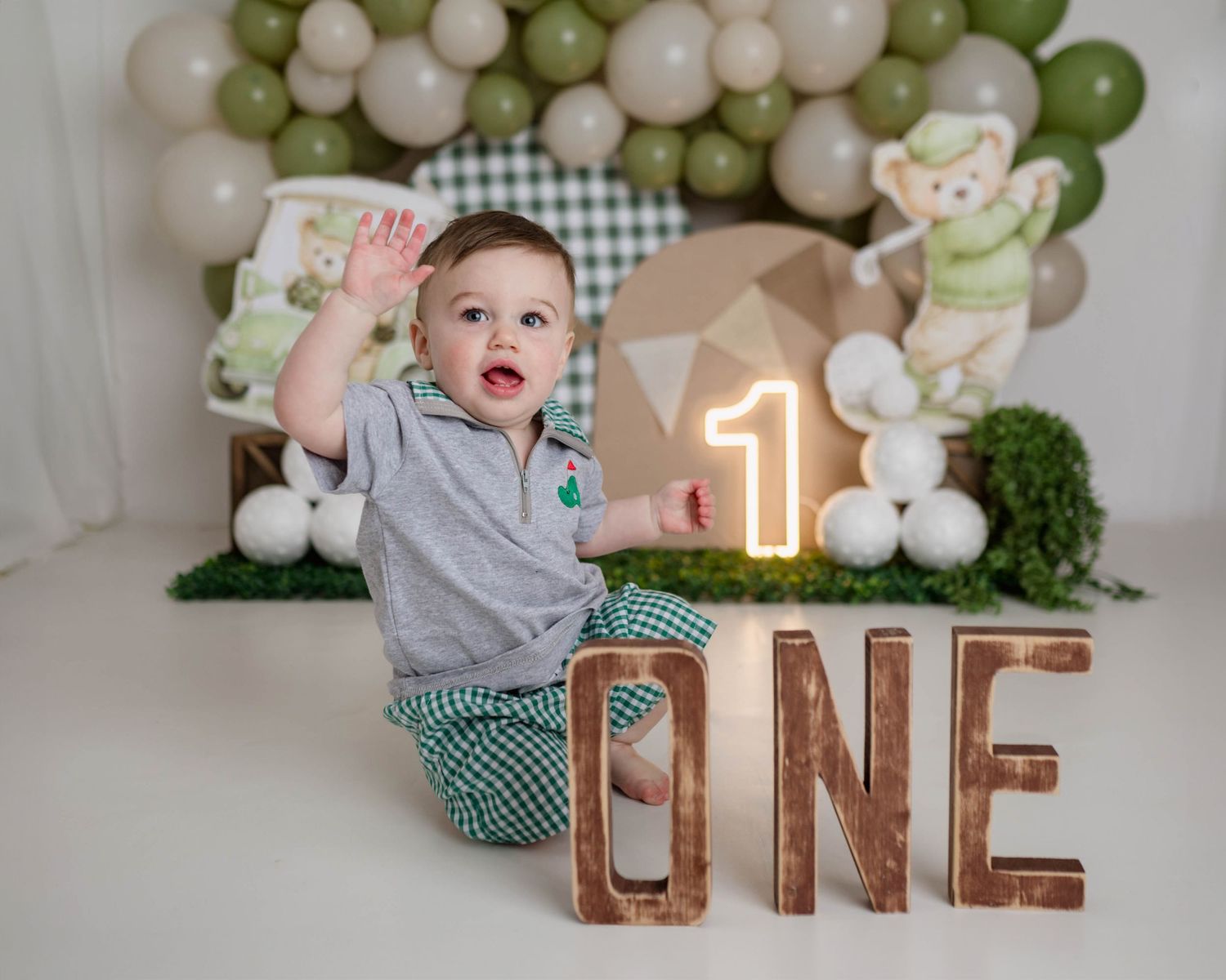 Baby boy celebrates first birthday with ONE sign, balloons, and golf theme decorations in professional photo shoot.