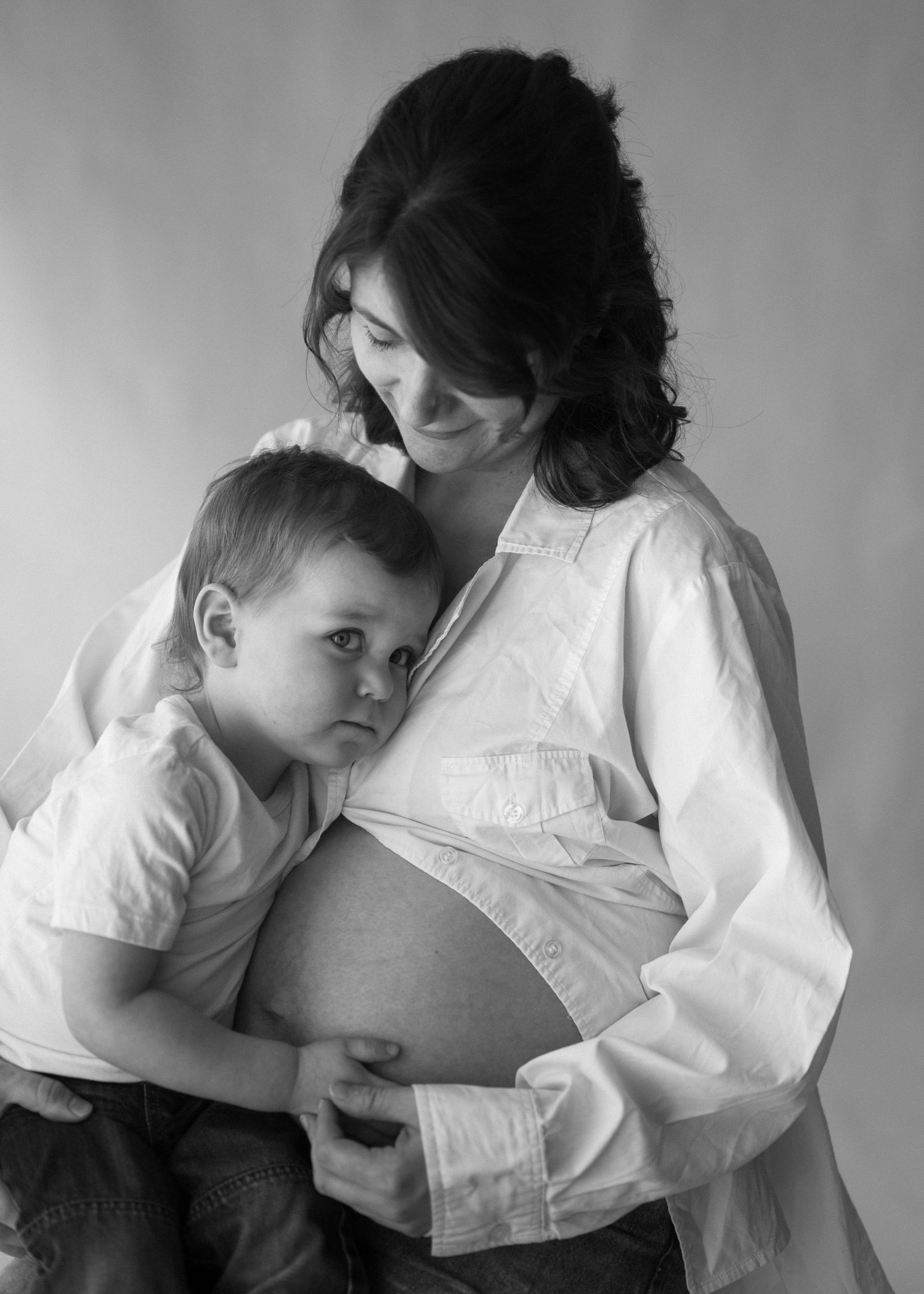 Black and white portrait of a pregnant mother and young son embracing her bare baby bump tenderly.