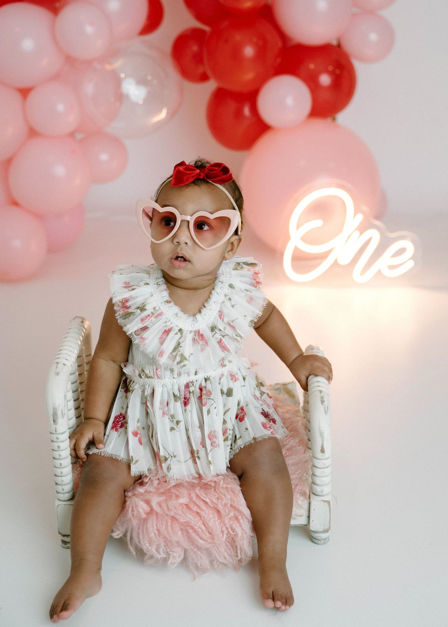 Baby girl in floral romper wears heart sunglasses on white chair beside glowing One neon sign.