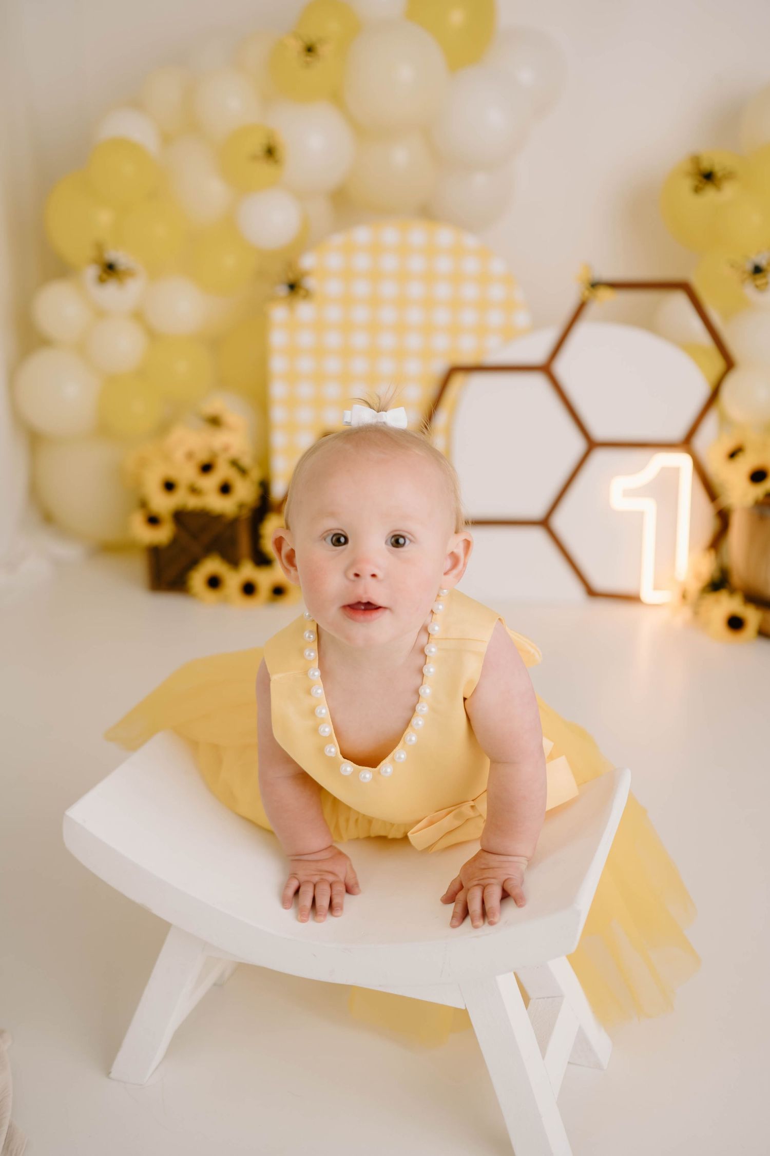 Baby girl in yellow dress at first birthday bee-themed party with honeycomb decor and yellow balloons.