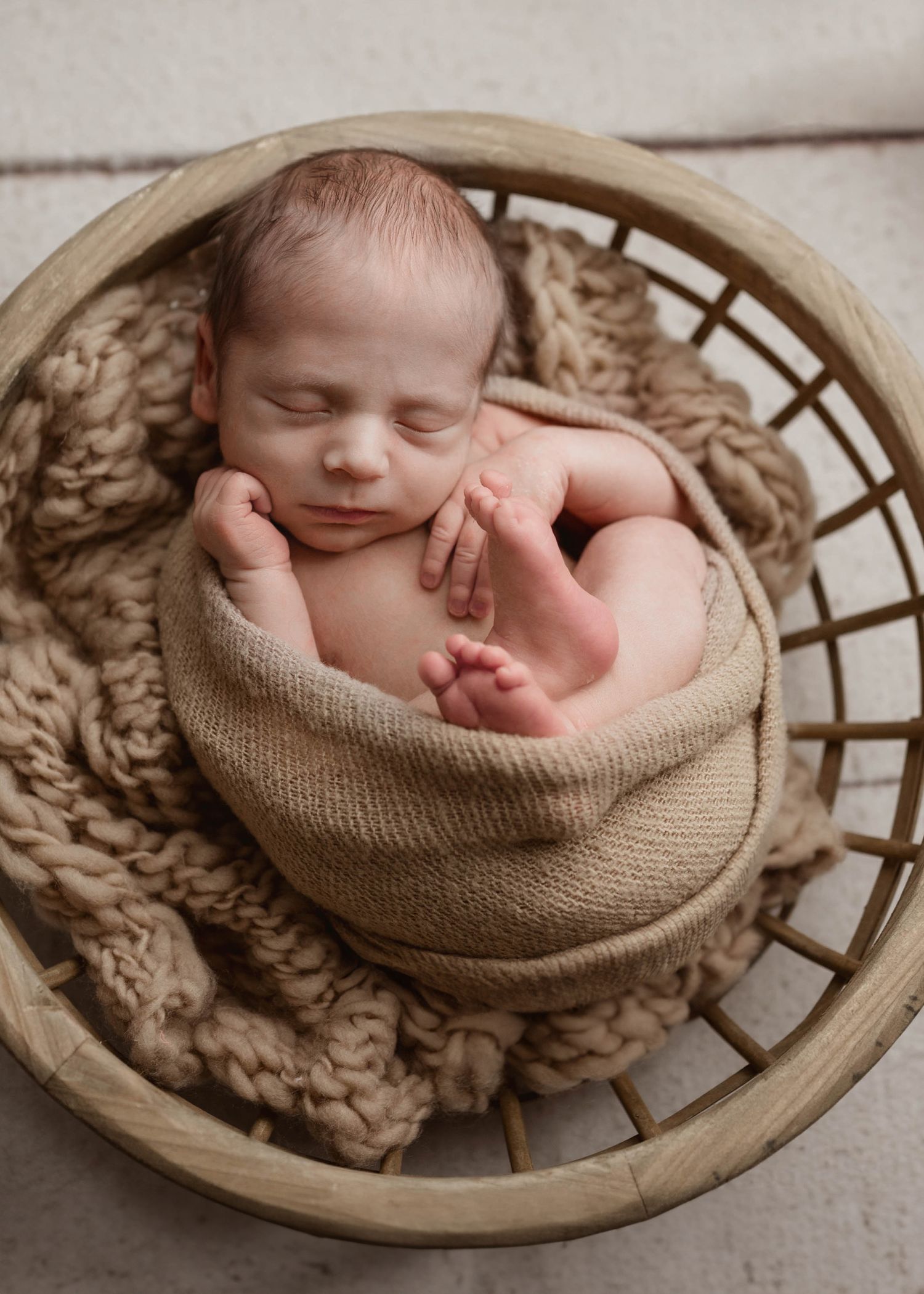 Sleeping newborn baby wrapped in tan swaddle nestled in a rustic wooden basket in Leduc Newborn Studio.