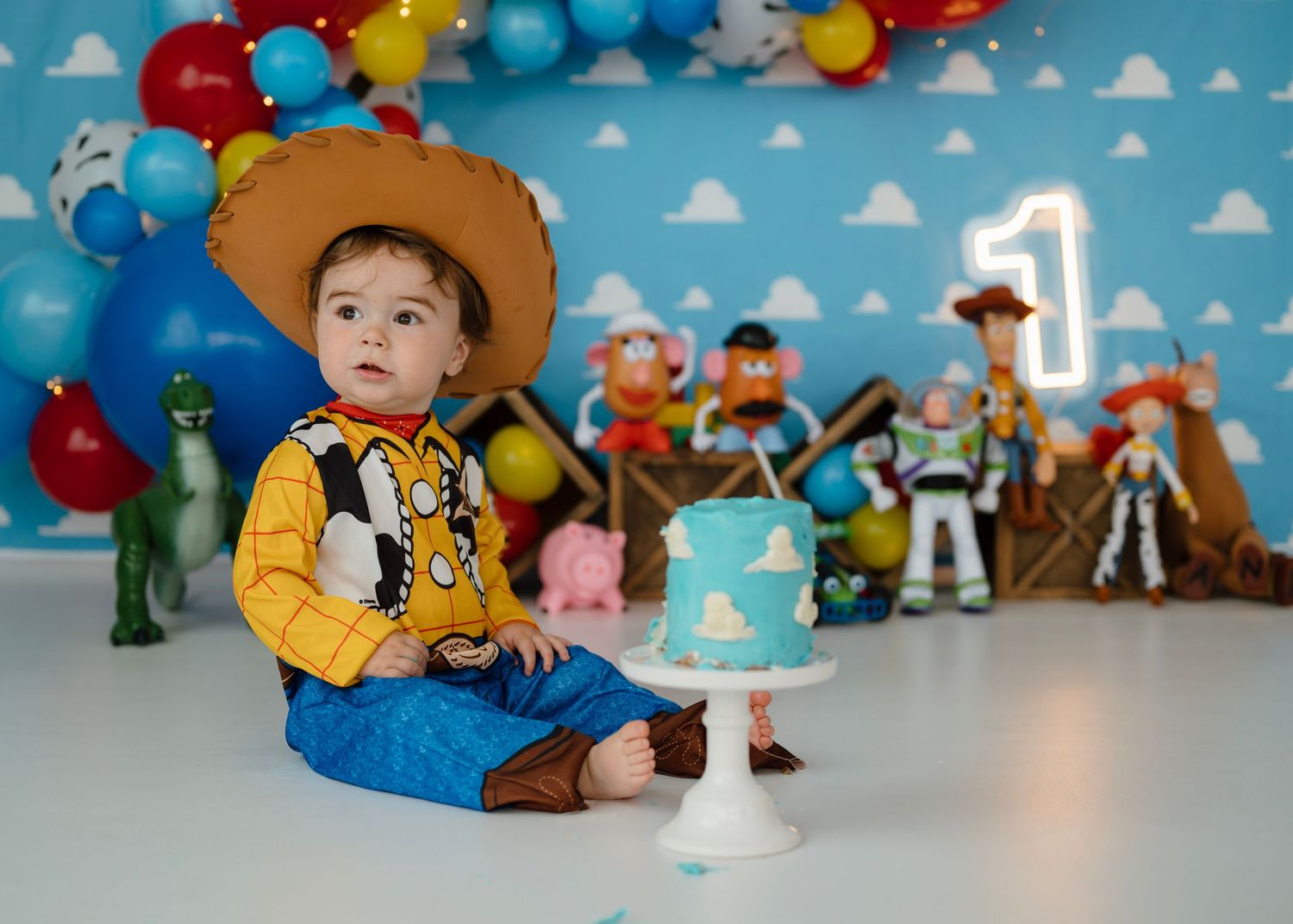 Baby boy in Woody costume at Toy Story themed first birthday cake smash with character toys.