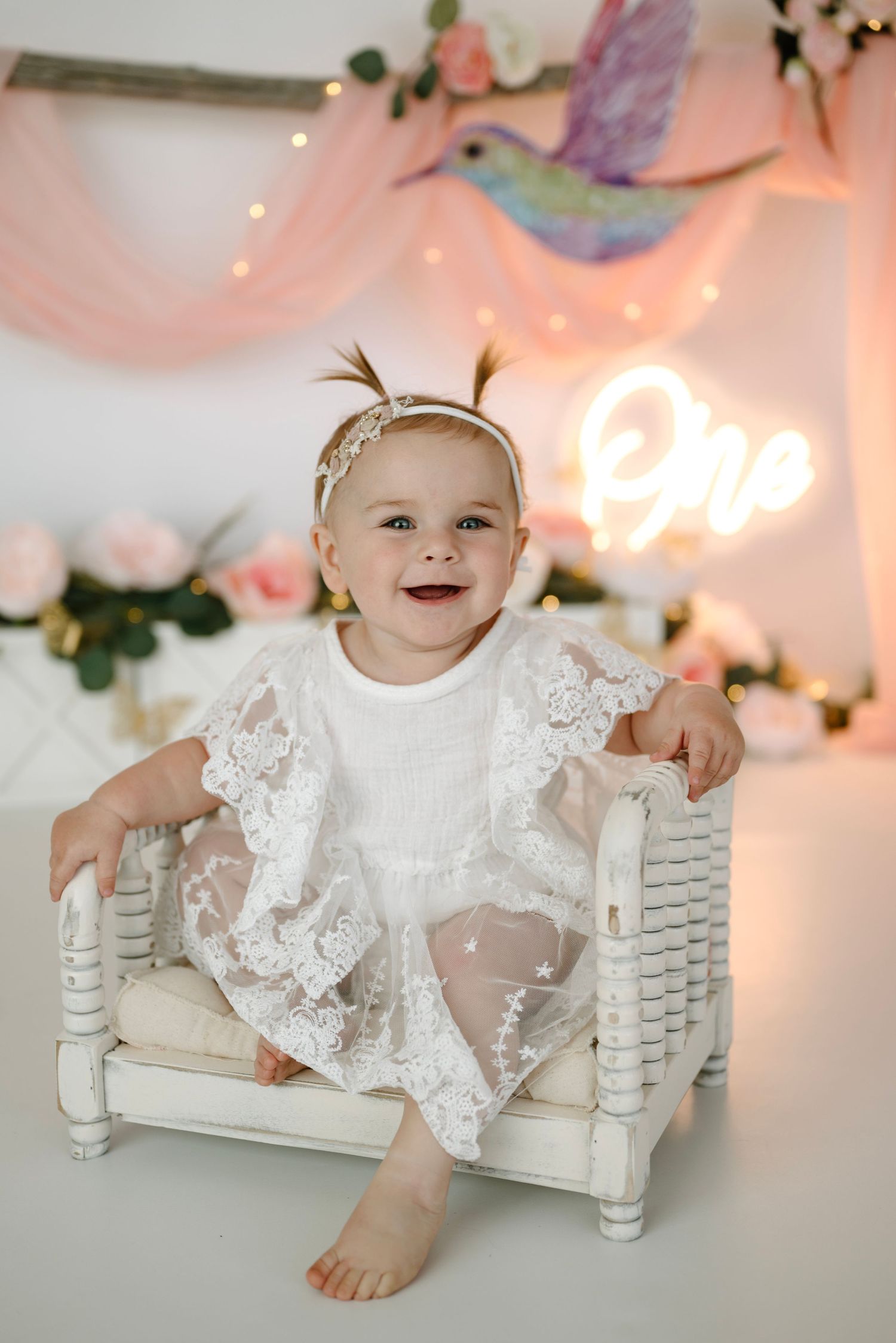 Smiling baby girl in white lace dress sitting on miniature bench with hummingbird birthday backdrop.