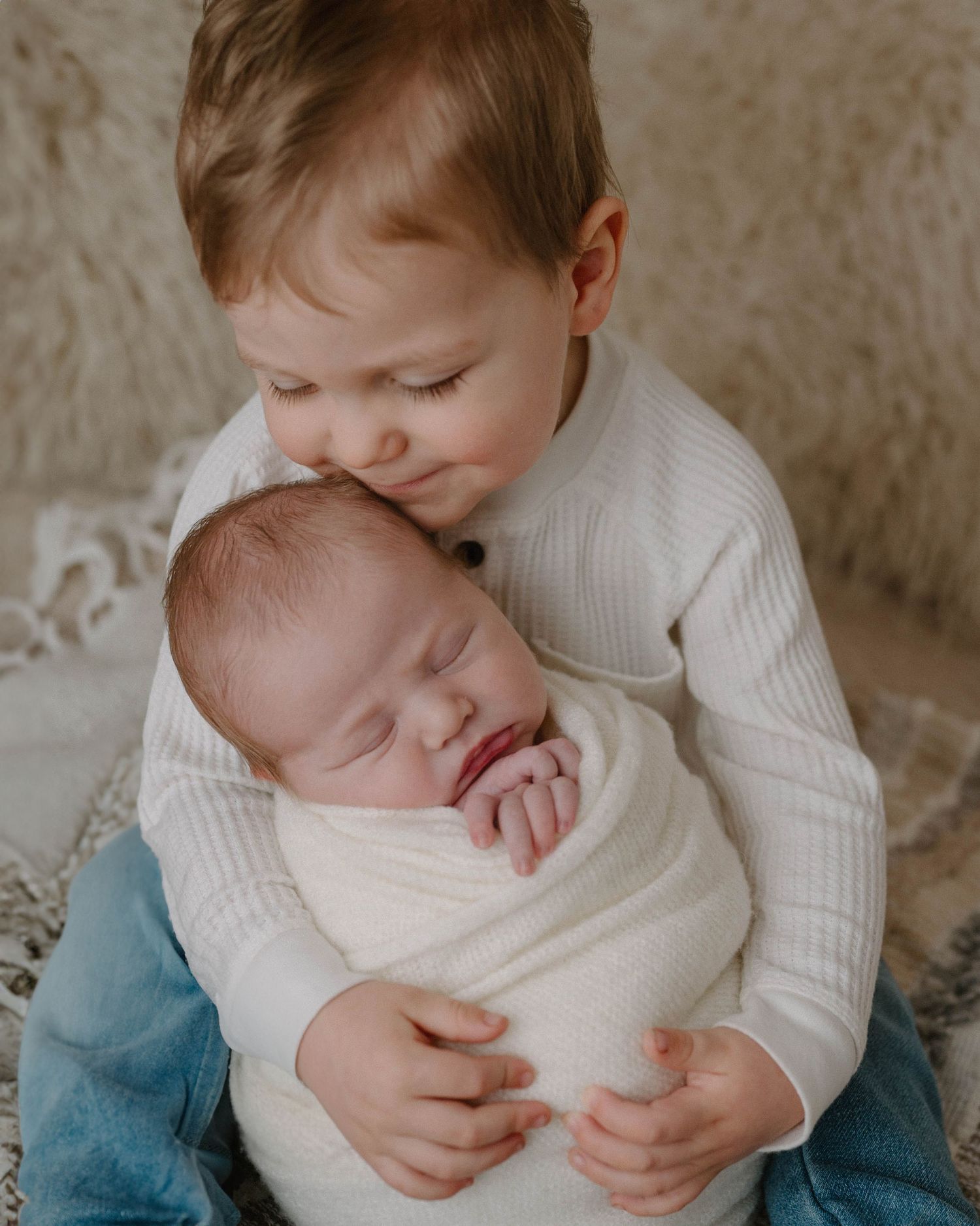 A toddler in a white sweater gently holds a sleeping newborn wrapped in a cream swaddle blanket.