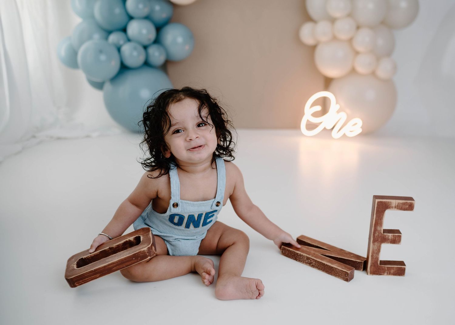 Smiling toddler in blue ONE romper sits on white floor with wooden letter props and balloon arch backdrop.