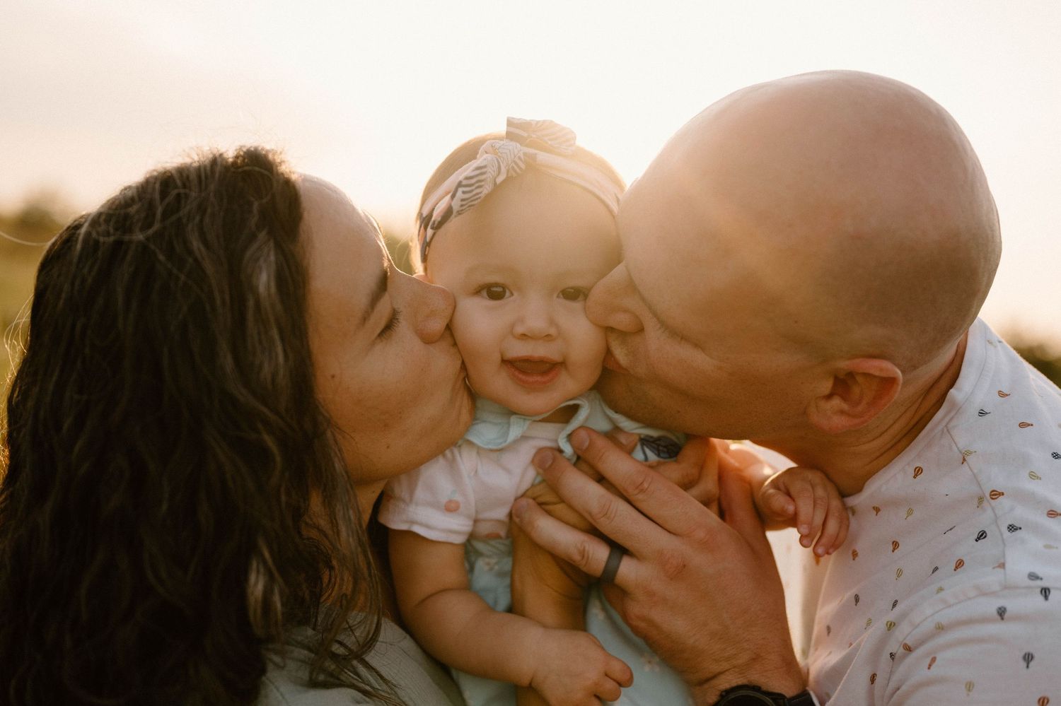 Loving parents share a tender moment kissing their smiling baby girl on both cheeks at sunset.