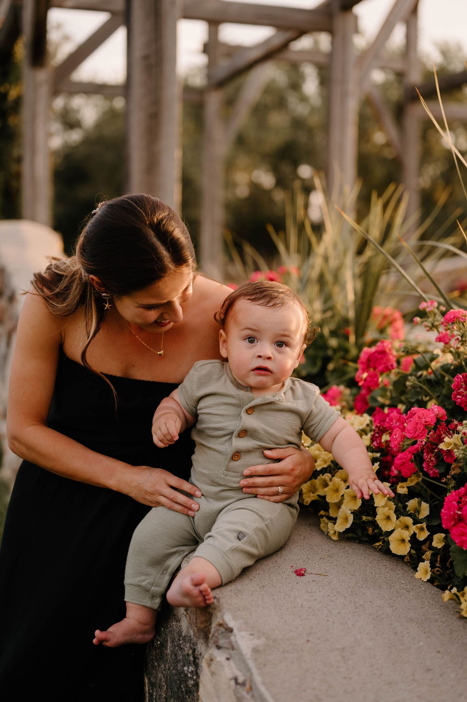 Mother in black dress holding baby boy in sage romper beside colorful flowers at golden hour at the Stone Barn Gardens in Leduc