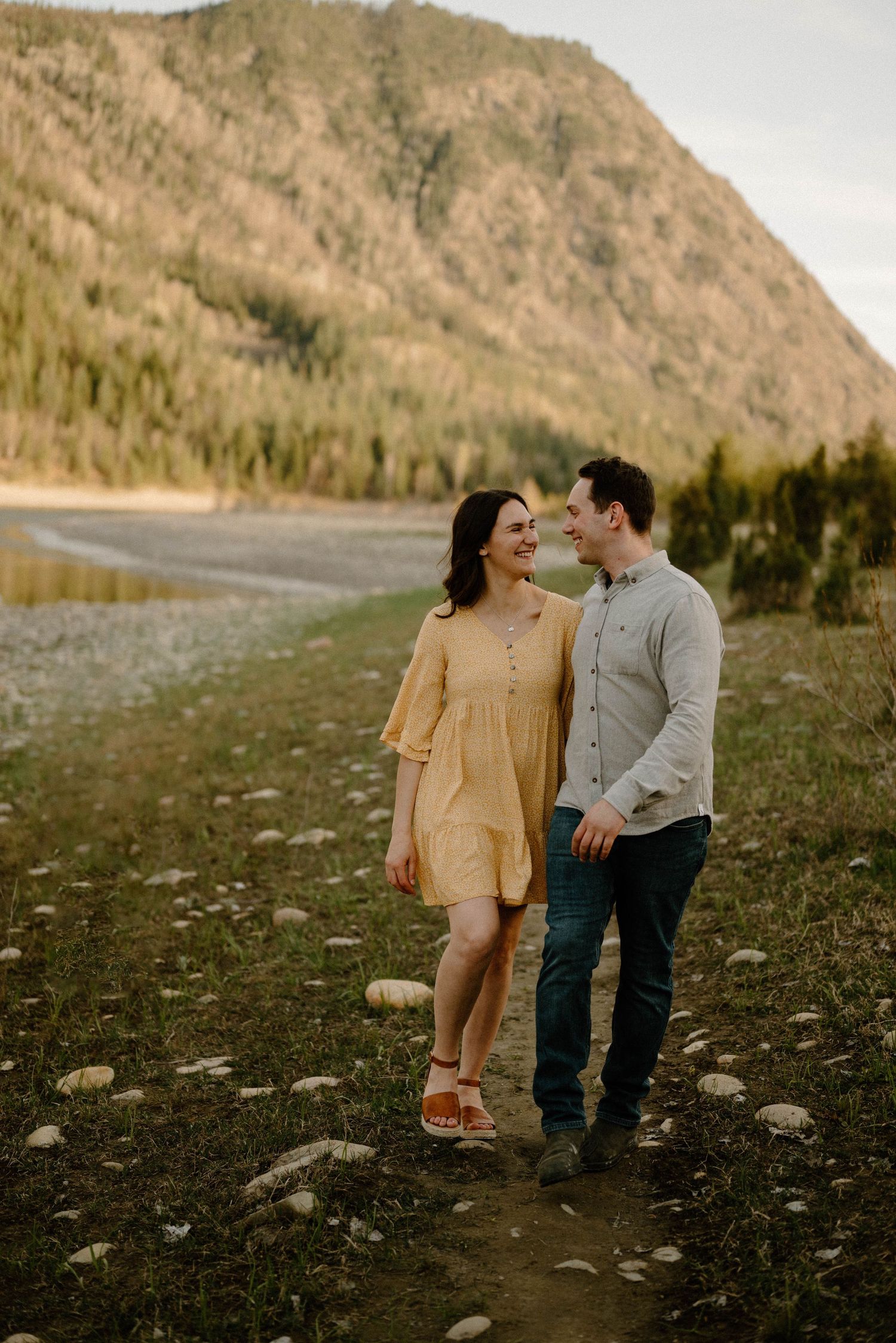Couple walking together smiling near a mountain lake at golden hour in casual attire.