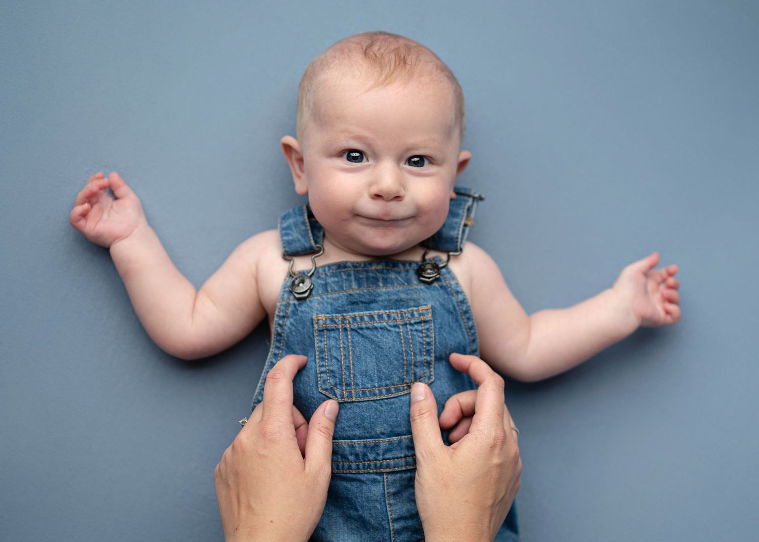 Smiling baby in denim overalls lying on a blue background with adult hands holding them.