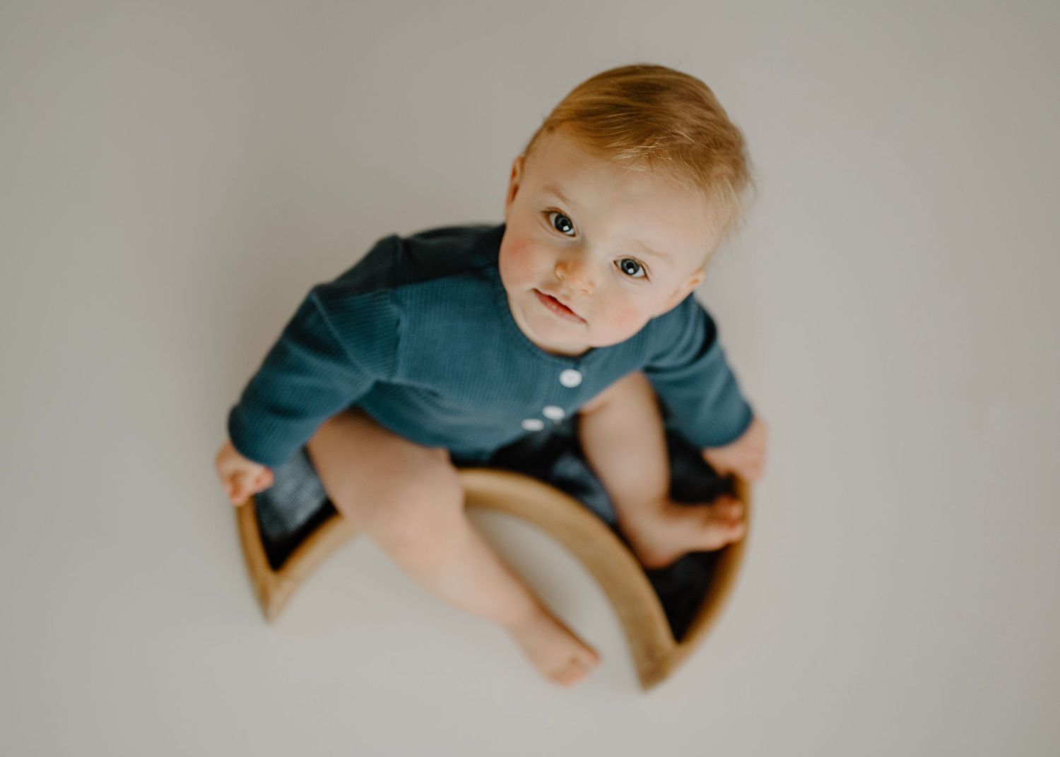 Adorable baby boy in blue romper looking up while seated on a wooden rocker chair.