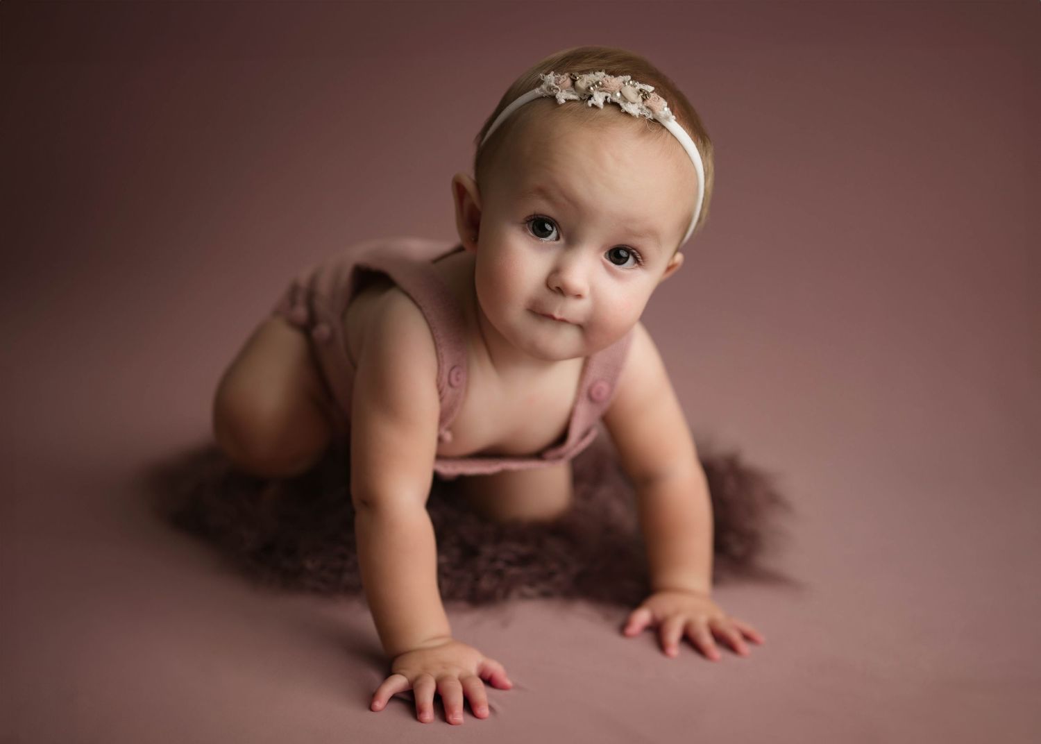Baby girl in pink romper and floral headband crawls on fluffy fur rug during studio portrait session.