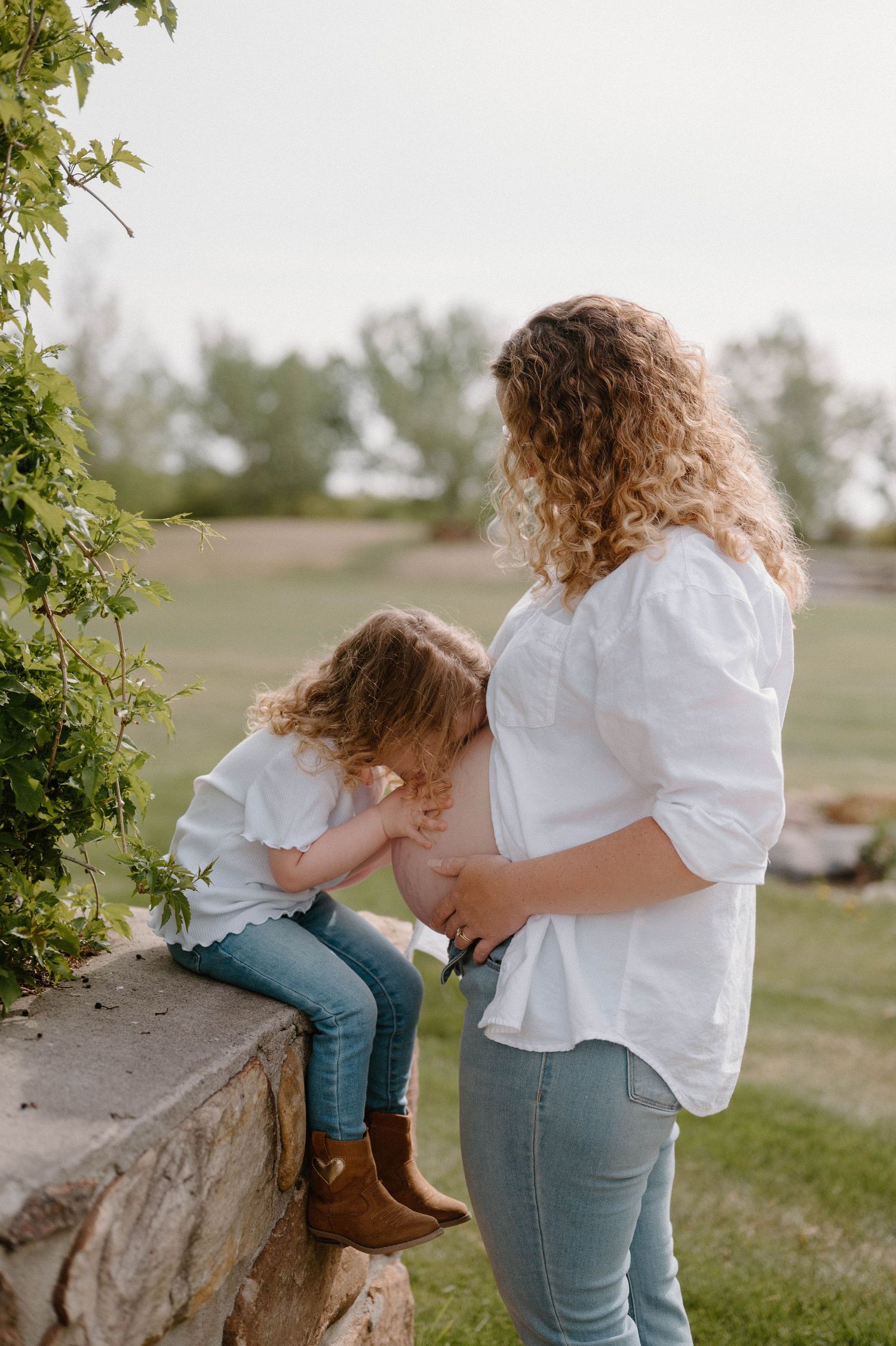 Pregnant mother in white shirt and jeans with curly hair, bonding with toddler daughter outdoors near stone wall.