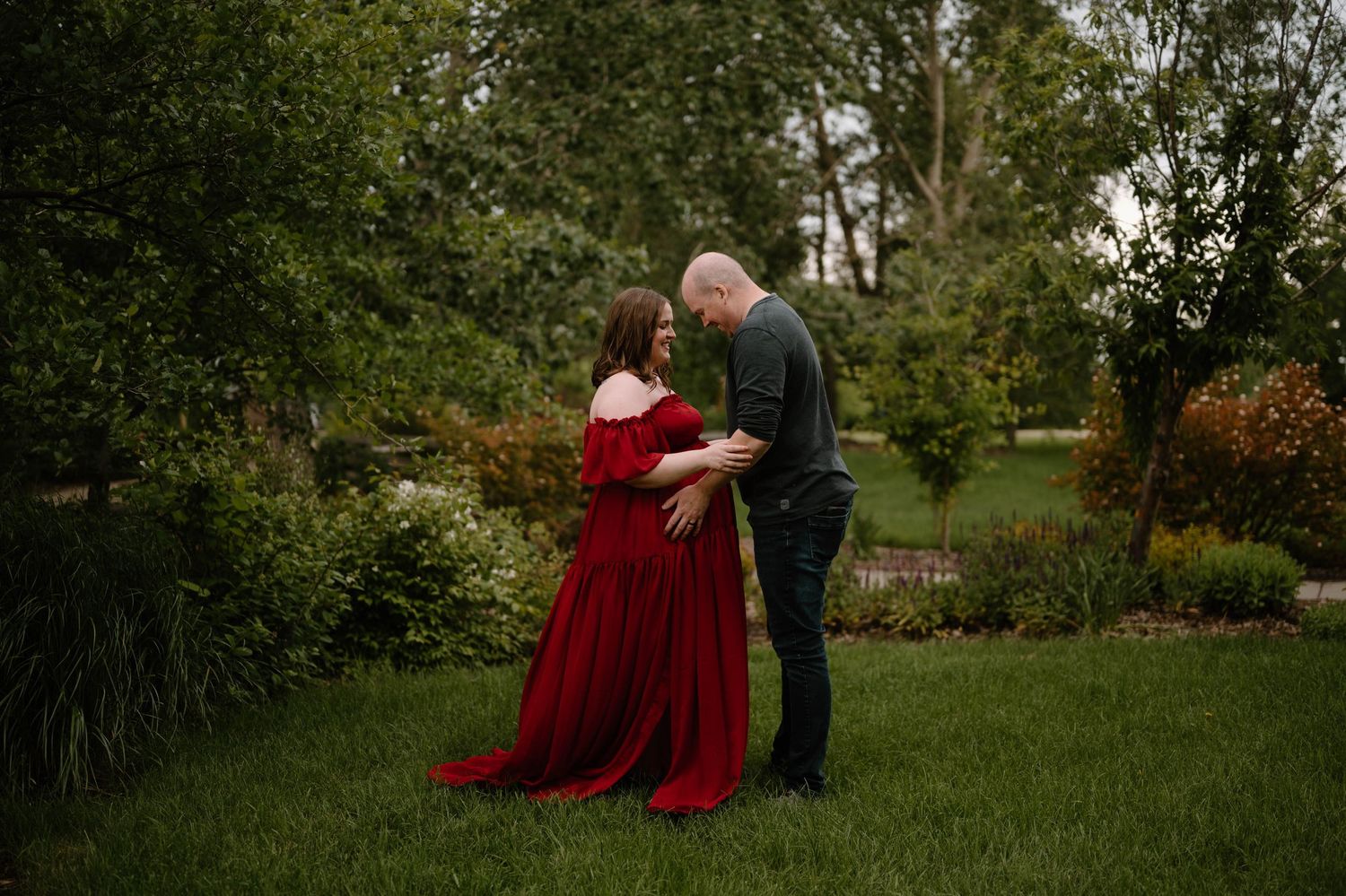 Couple sharing a tender moment in a lush garden, woman in flowing red maternity gown, man in dark shirt.