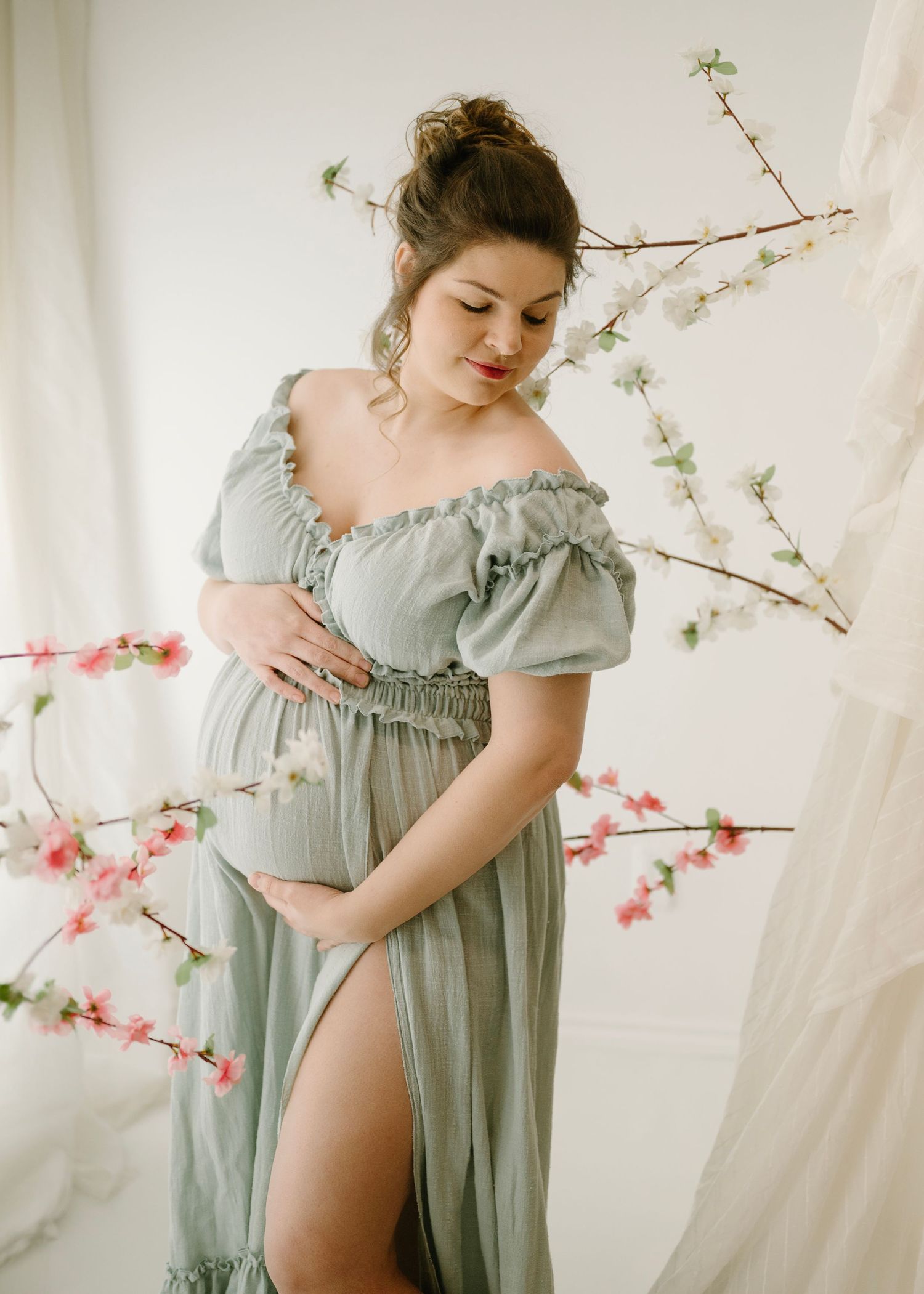 Pregnant woman in sage green off-shoulder maternity gown cradles belly beside pink and white cherry blossoms.