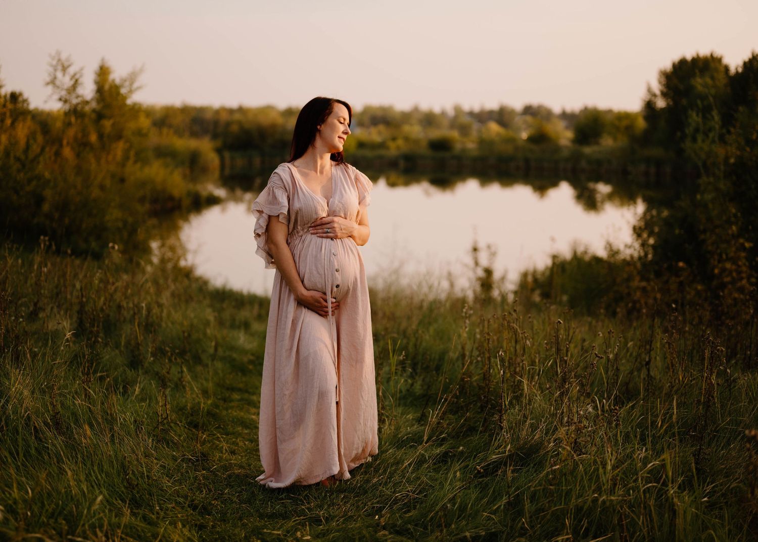 Pregnant woman in flowing pink dress standing by a calm lake at golden hour.