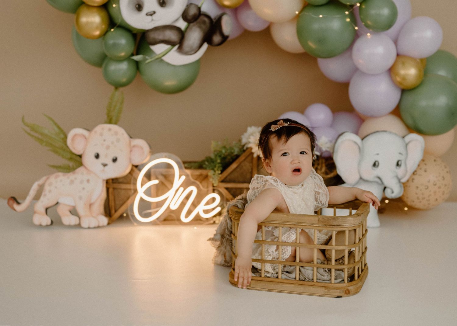 Baby girl stands in wicker basket beside ONE neon sign with jungle animal decor.