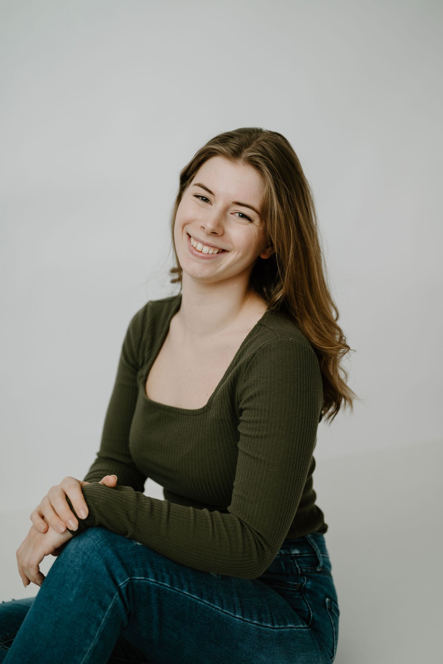 Smiling young brunette woman in olive green top and jeans sitting casually on white background.